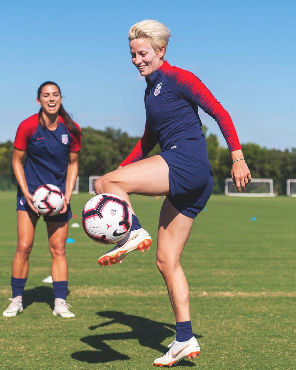 Alex Morgan 桌布 壁紙 — 兩名微笑的女足球員身穿藍色和紅色球衣，在陽光明媚的草地上訓練，其中一人凌空踢球。