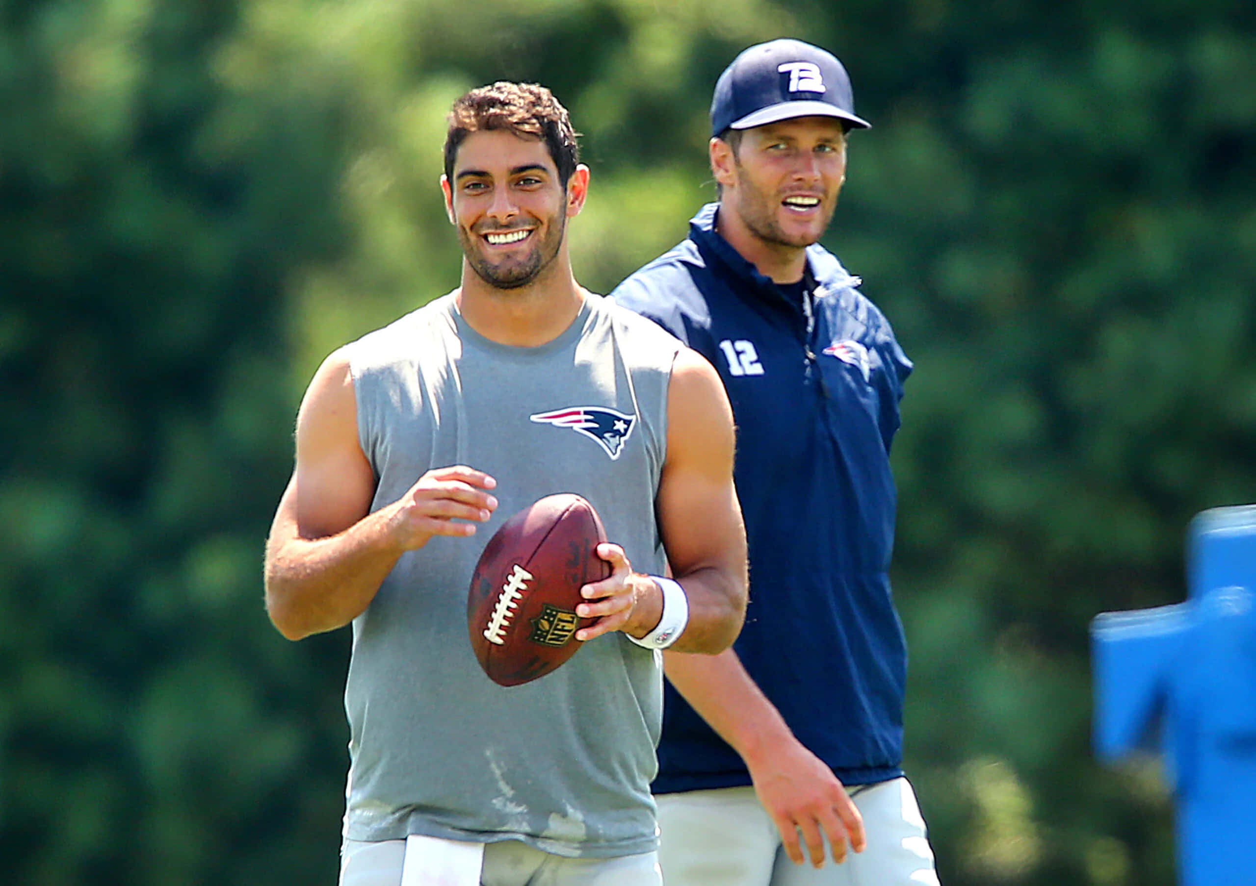 Jimmy Garoppolo 桌布 壁紙 — 兩名微笑的美式足球員身穿新英格蘭愛國者隊裝備，其中一人在陽光明媚的日子裡在戶外拿著球。