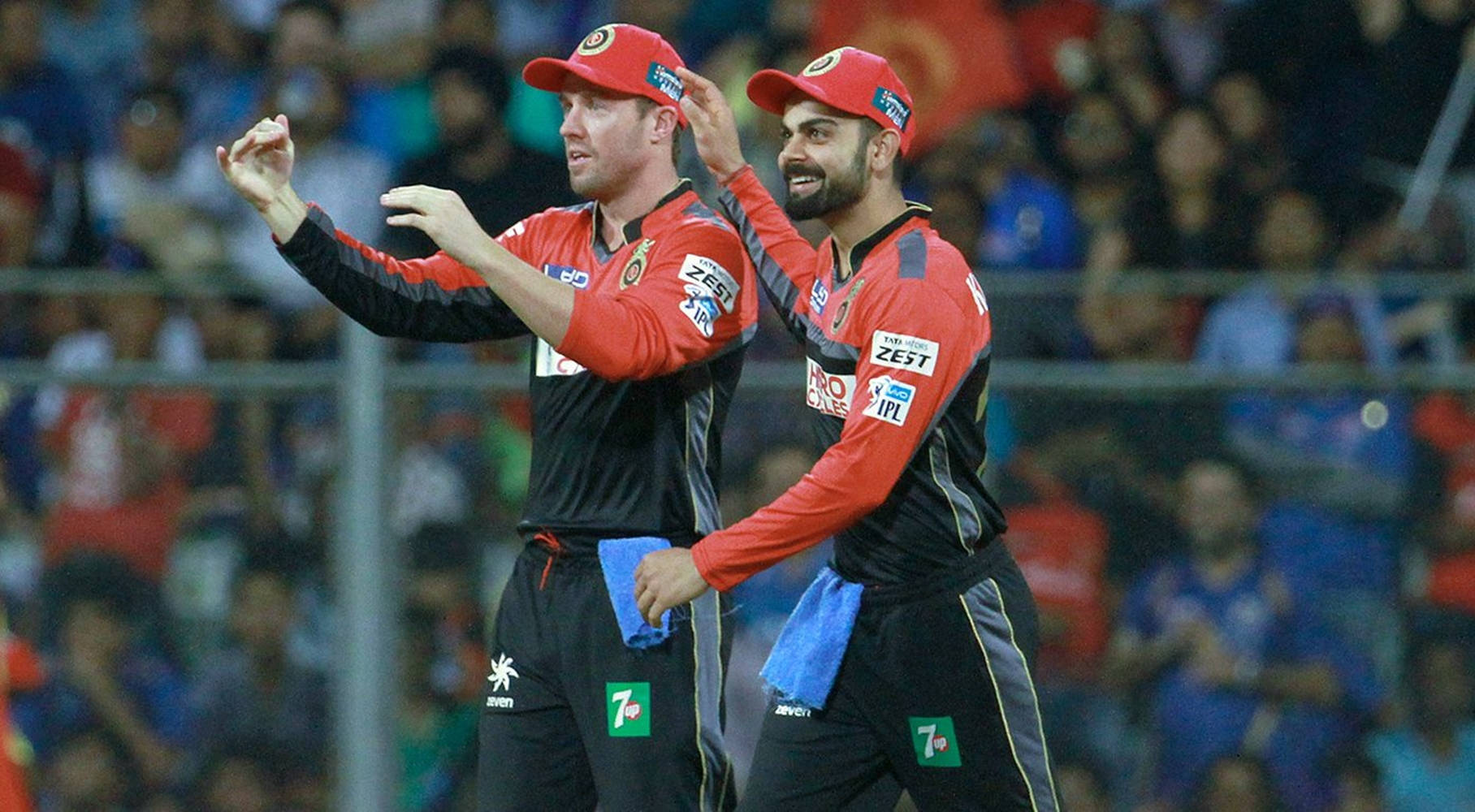 virat kohli ipl 桌布 壁紙 — 兩名戴紅色帽子和穿著紅色球衣的板球運動員在燈光明亮的體育場上一起慶祝，背景是模糊的球迷。