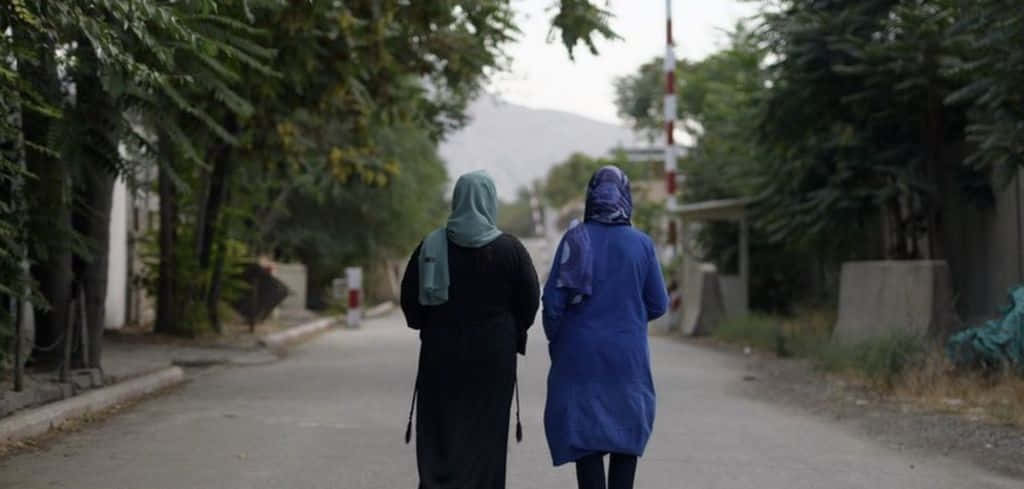 Taliban 桌布 壁紙 — 兩名戴頭巾的女子沿著綠樹成蔭的街道走遠，朝向遠方的山丘，天空多雲。