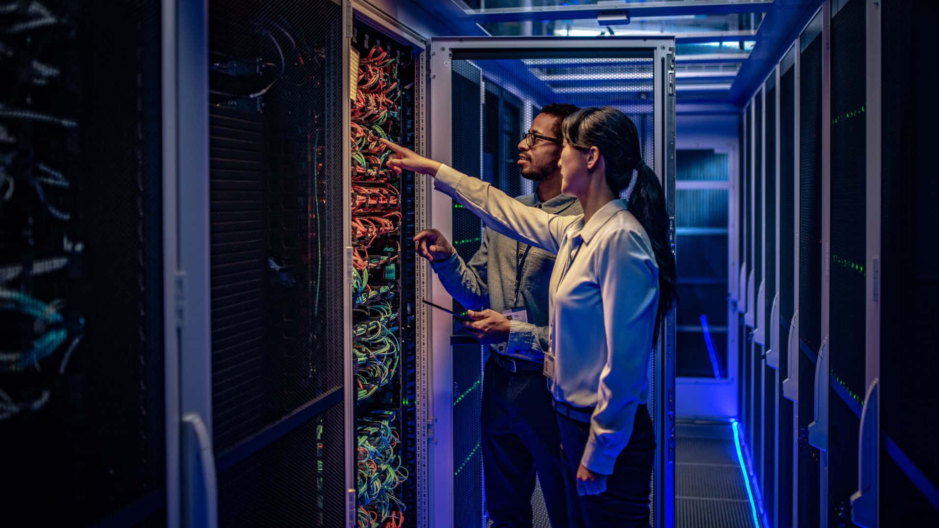 Gaming Servers 桌布 壁紙 — 兩名技術人員在昏暗的數據中心檢查發光的伺服器機架，沐浴在藍光中。