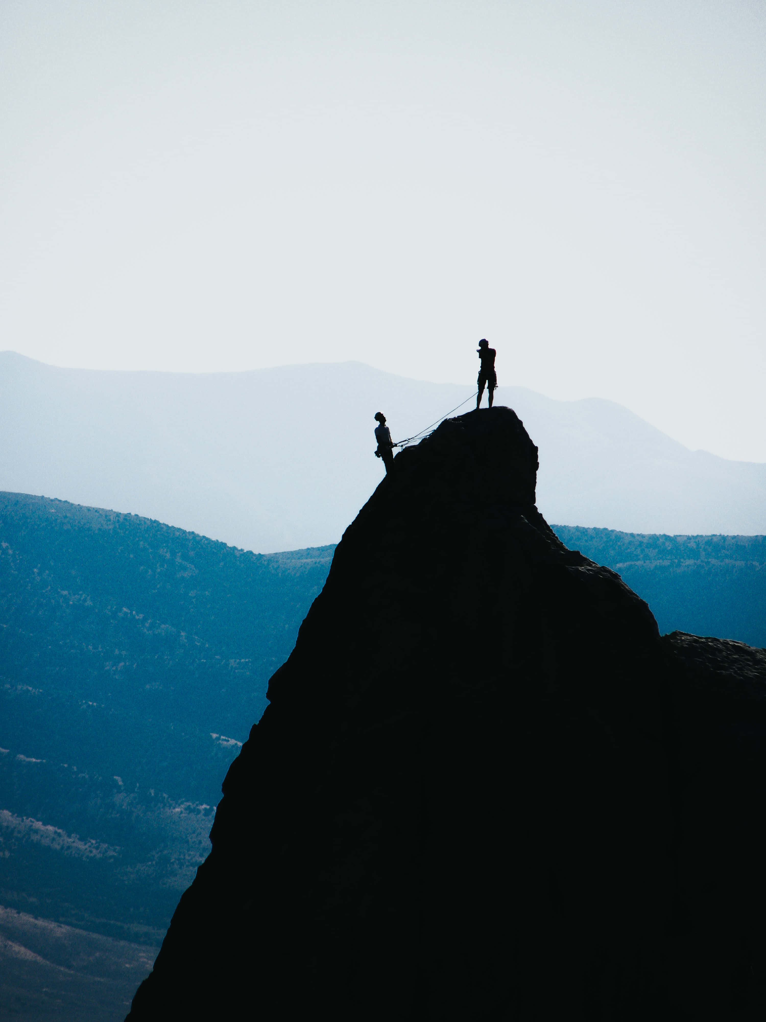 Silhouette 桌布 壁紙 — 兩名攀岩者嘅剪影企喺鋸齒狀山峰上，背景係柔和日光下嘅朦朧藍色山脈。