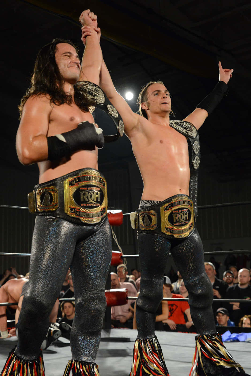 Caption: American Professional Wrestler Matt Jackson in Action 壁紙 — 兩名汗流浹背的摔角手身穿黑色閃亮緊身褲和ROH冠軍腰帶，在擂台上高舉雙臂慶祝勝利。