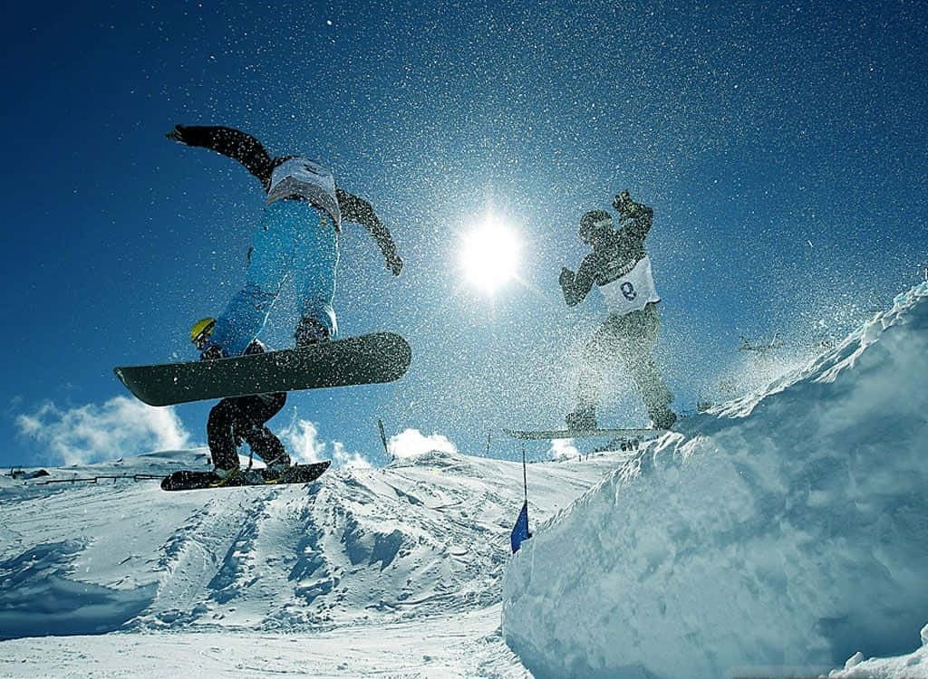 Winter Games 桌布 壁紙 — 兩名滑雪板運動員在白雪覆蓋的山坡上，迎著熾熱的太陽，在清爽的冬日空氣中翱翔。