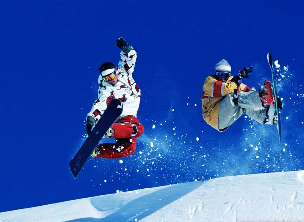 Winter Sports 桌布 壁紙 — 兩名滑雪板運動員在雪坡上空飛過清澈的藍天，噴灑著雪粒。