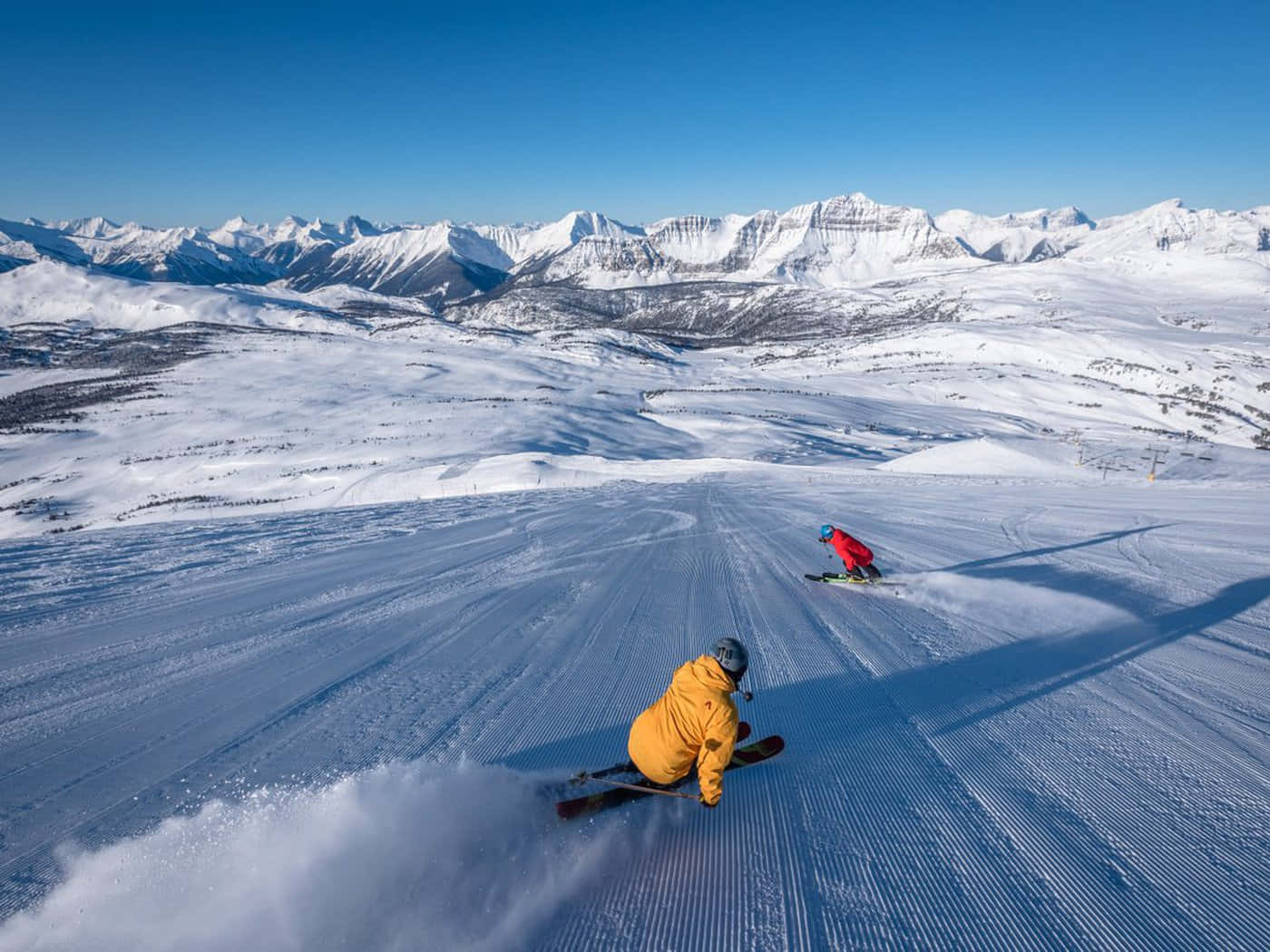 Skiing 桌布 壁紙 — 兩名滑雪者喺陽光照耀嘅修整雪道上 carve 落嚟，身後雪花四濺，背景係晴朗藍天下遠處嘅雪山。