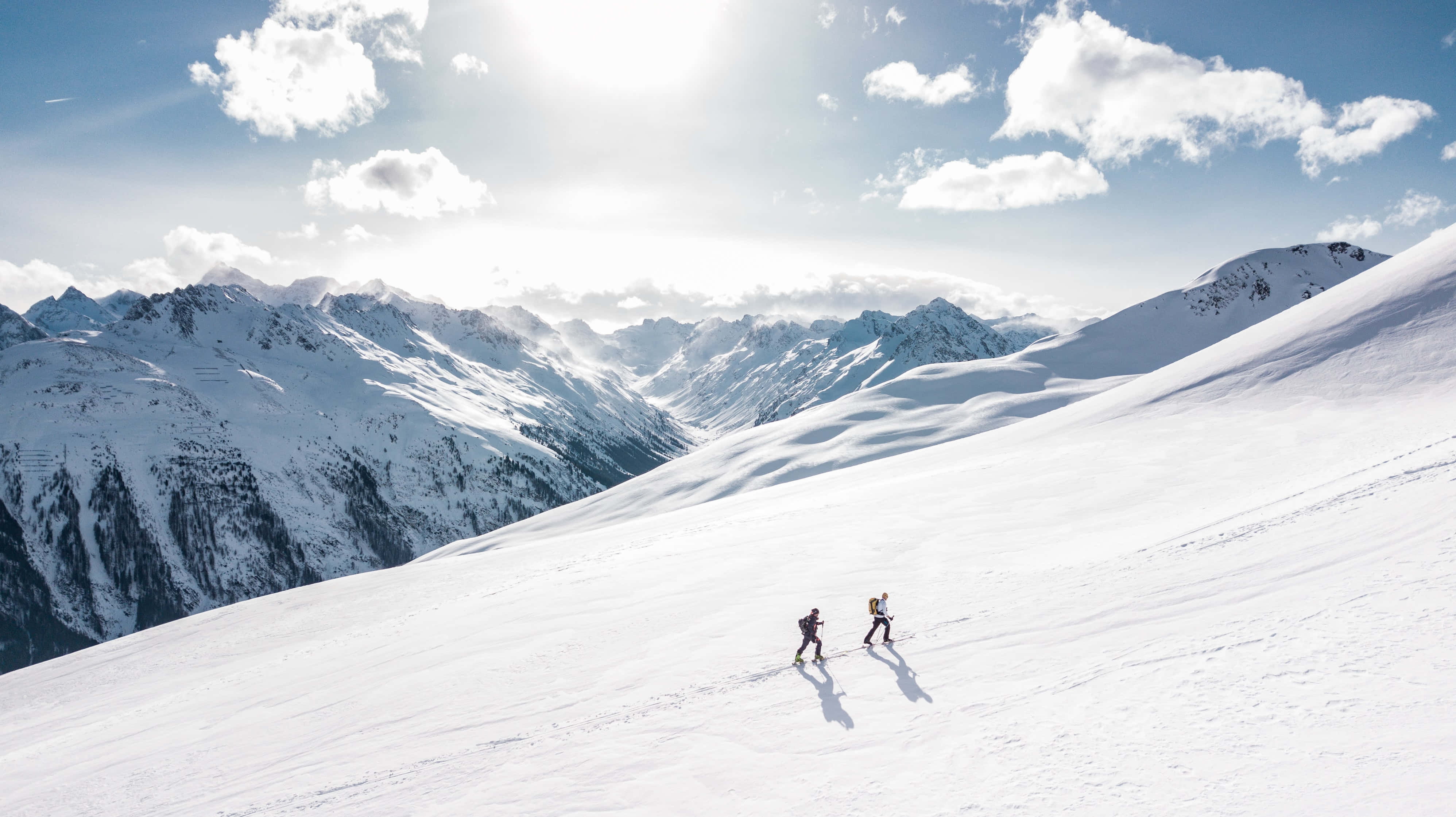 寒冷 桌布 壁紙 — 兩名滑雪者在一片廣闊、陽光普照的積雪山坡上滑行，遠處有鋸齒狀的山峰和散落的雲朵。