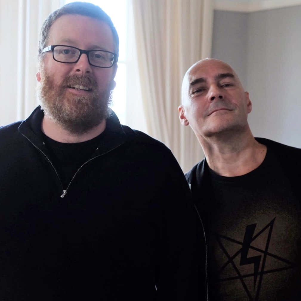 Grant Morrison 桌布 壁紙 — 兩名男子在室內靠近窗戶站立，一人戴眼鏡留鬍鬚，另一人禿頭，兩人都穿著黑色衣服。