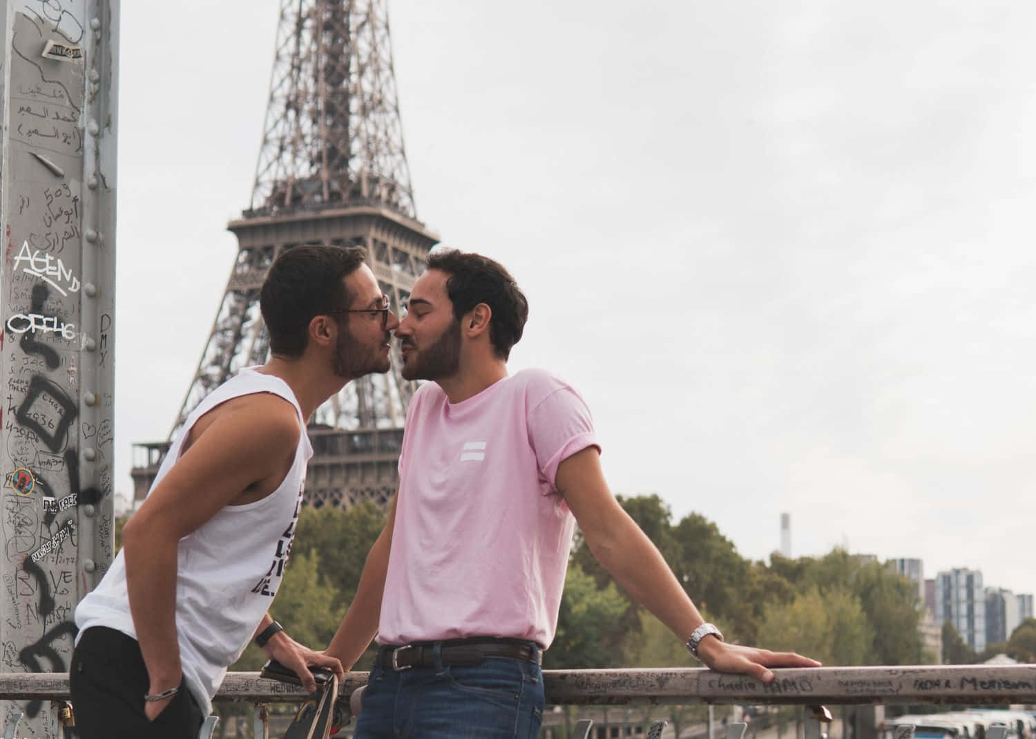 french 桌布 壁紙 — 兩名男子在巴黎的一座橋上溫柔地接吻，艾菲爾鐵塔在陰天的天空下隱約出現在他們身後。
