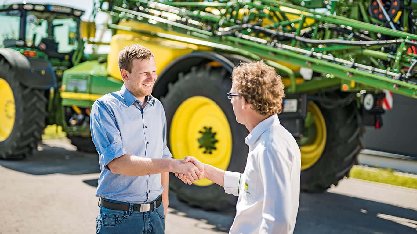 John Deere 桌布 壁紙 — 兩名男子在明亮的陽光下，在大型綠色和黃色農業機械前握手。