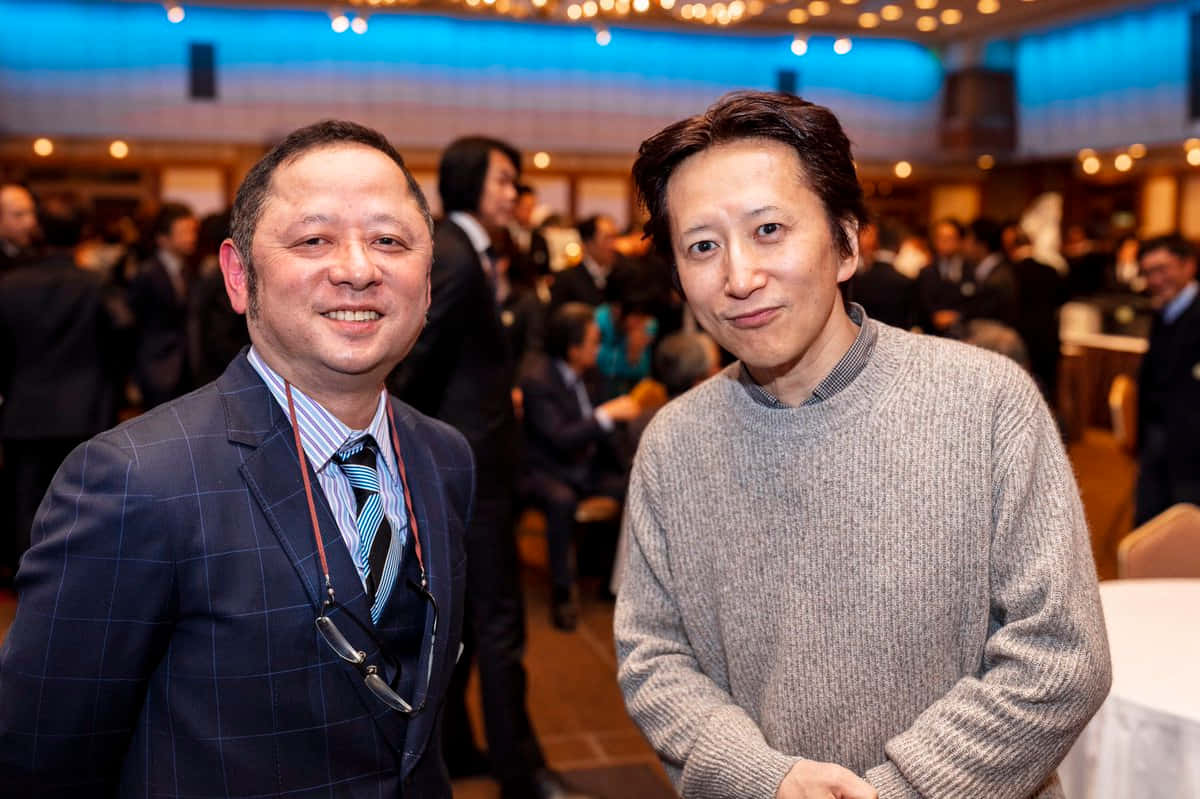 Hirohiko Araki 桌布 壁紙 — 兩名男子在溫暖的環境燈光下，於擁擠的室內活動中對著鏡頭微笑。