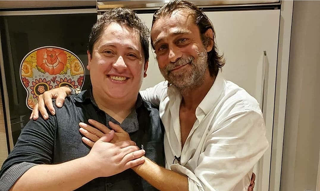 Jordi Mollà 桌布 壁紙 — 兩名男子在舒適的室內環境中熱情擁抱，身後是色彩繽紛的糖骷髏藝術品。