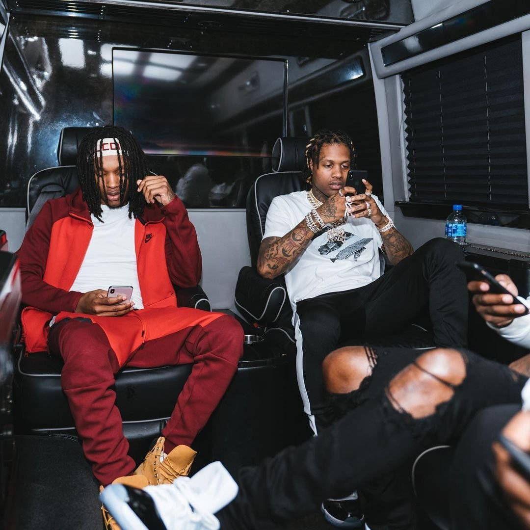 King Von And Lil Durk 桌布 壁紙 — 兩名男子在豪華客貨車內休息，一人身穿紅色運動服，兩人都專注於手機，紋身可見，皮革座椅，環境照明。