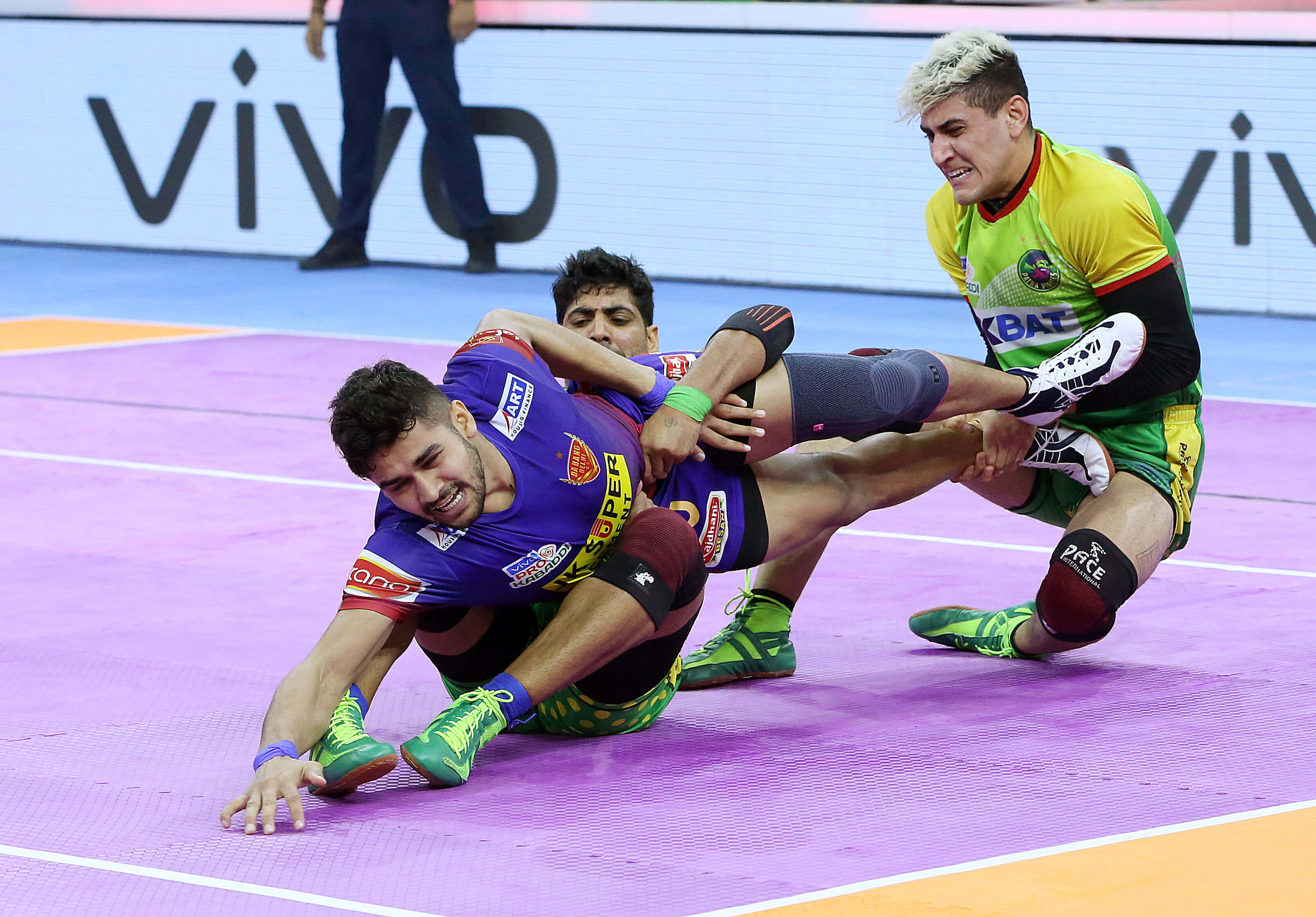 Kabaddi 桌布 壁紙 — 兩名男性摔角手在明亮的場館燈光下，於充滿活力的紫色墊子上激烈纏鬥，進行一場競爭激烈的比賽。
