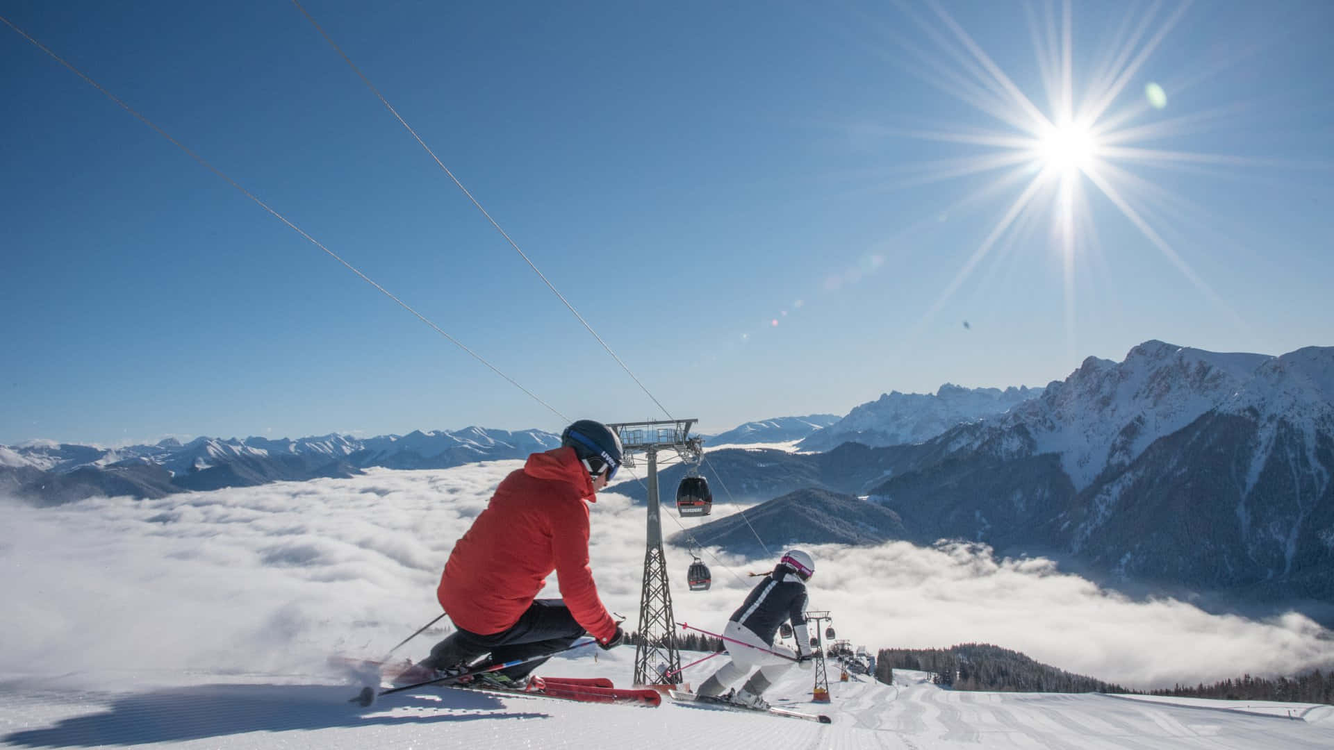 Skiing 桌布 壁紙 — 兩名穿著鮮豔裝備的滑雪者，在熾熱的陽光下，停留在雲海之上白雪覆蓋的山頂。