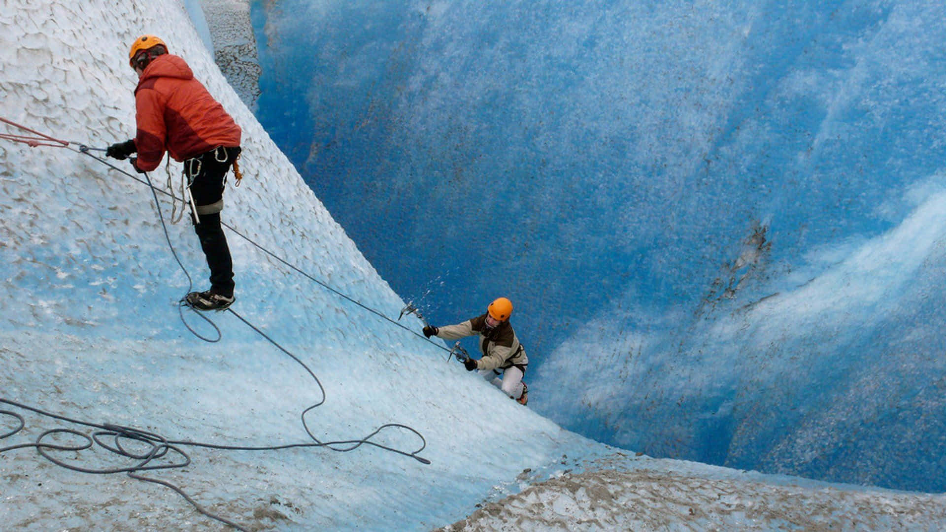 Ice Climbing 桌布 壁紙 — 兩名穿著鮮豔裝備的登山者用繩索和冰斧攀登陡峭的冰藍色冰川面。