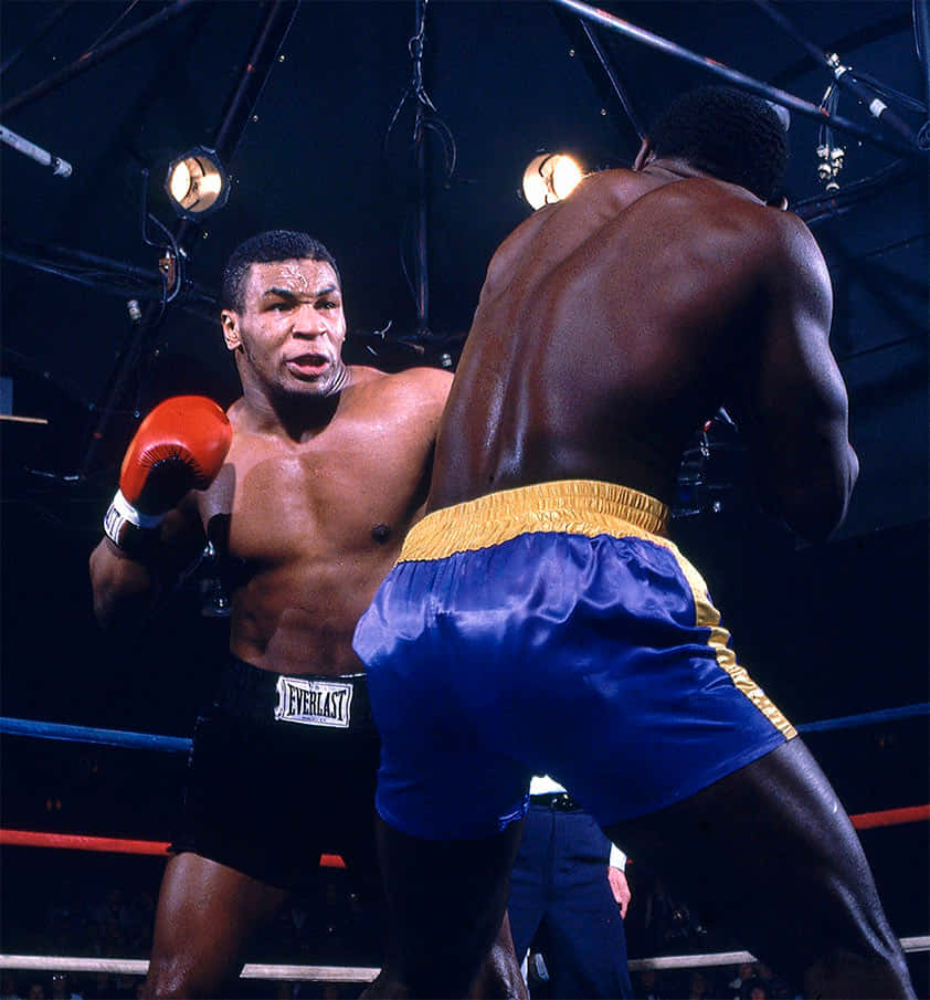 mike tyson 桌布 壁紙 — 兩名肌肉發達的拳擊手在昏暗的擂台上，在明亮的聚光燈下交鋒，一人穿著黑色短褲，另一人穿著藍色和金色短褲。