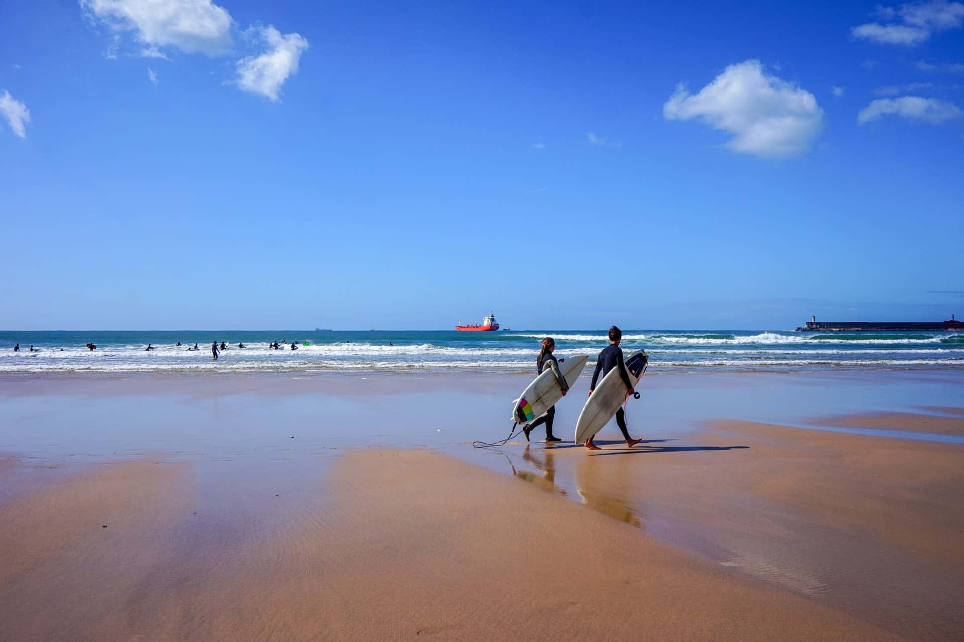 Portuguese Beach 桌布 壁紙 — 兩名衝浪者在明亮的藍天下，帶著衝浪板走過濕沙，天空有散落的雲朵和遠處的海浪。