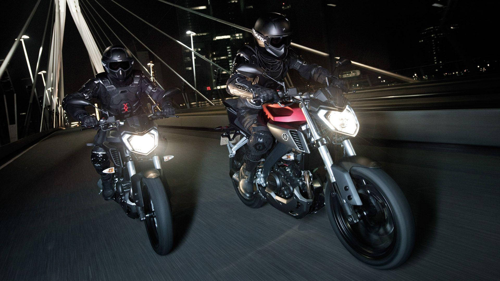 yamaha mt 15 桌布 壁紙 — 兩名身穿全黑裝備的電單車手在夜間於一座現代化橋樑上競速，身後城市燈光閃爍。