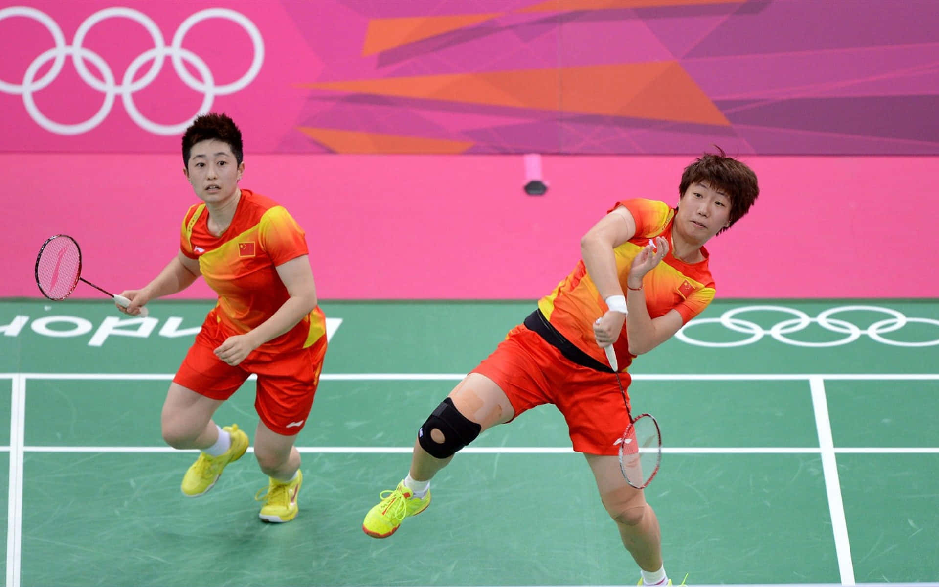 Badminton 桌布 壁紙 — 兩名身穿橙色球衣的羽毛球運動員在明亮的場館燈光下，於綠色球場上激烈對戰。