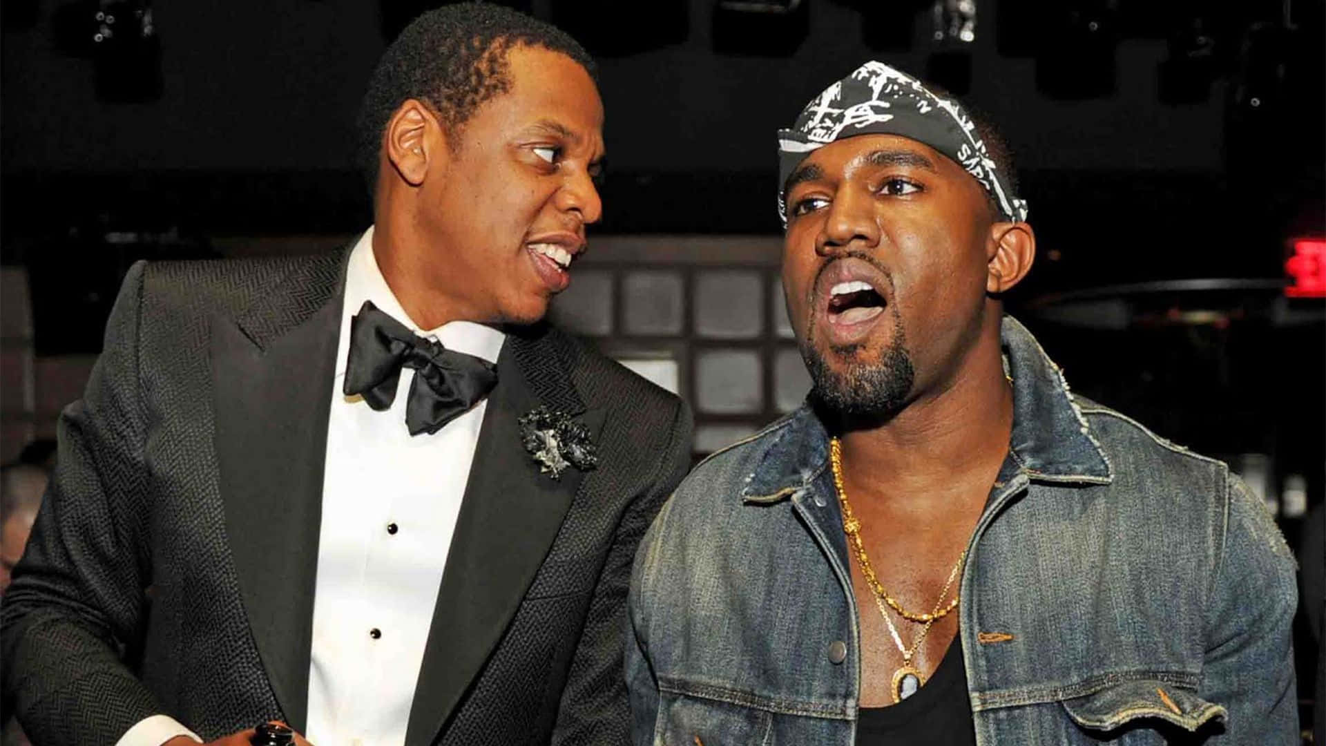 Kanye 桌布 壁紙 — 兩名身穿正式及休閒服裝的男子在昏暗的活動燈光下進行熱烈交談。