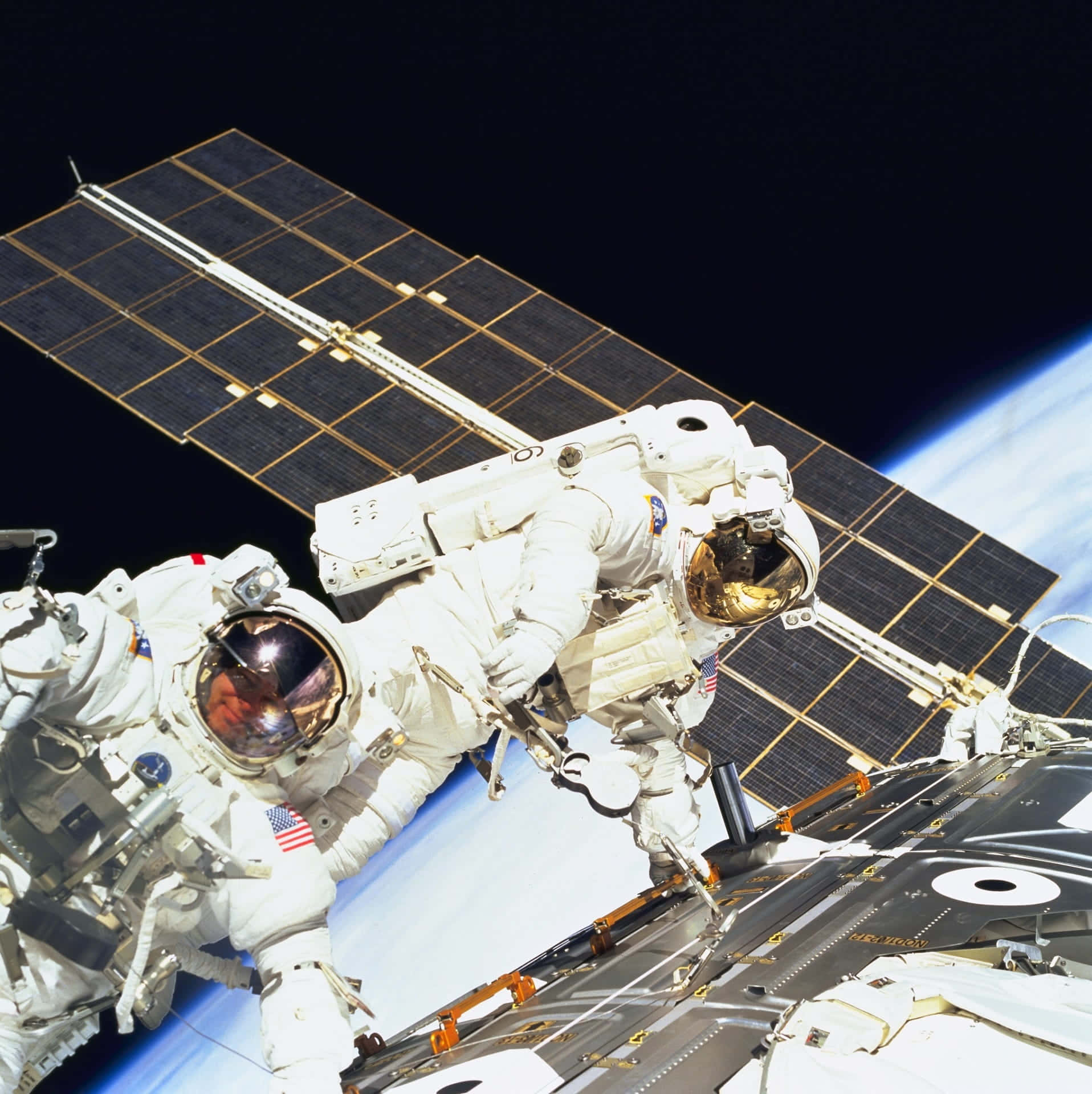 Space Mission 桌布 壁紙 — 兩名身穿白色太空衣的太空人在衛星外進行太空漫步，背景是黑色虛空和地球彎曲的地平線，衛星帶有太陽能板。