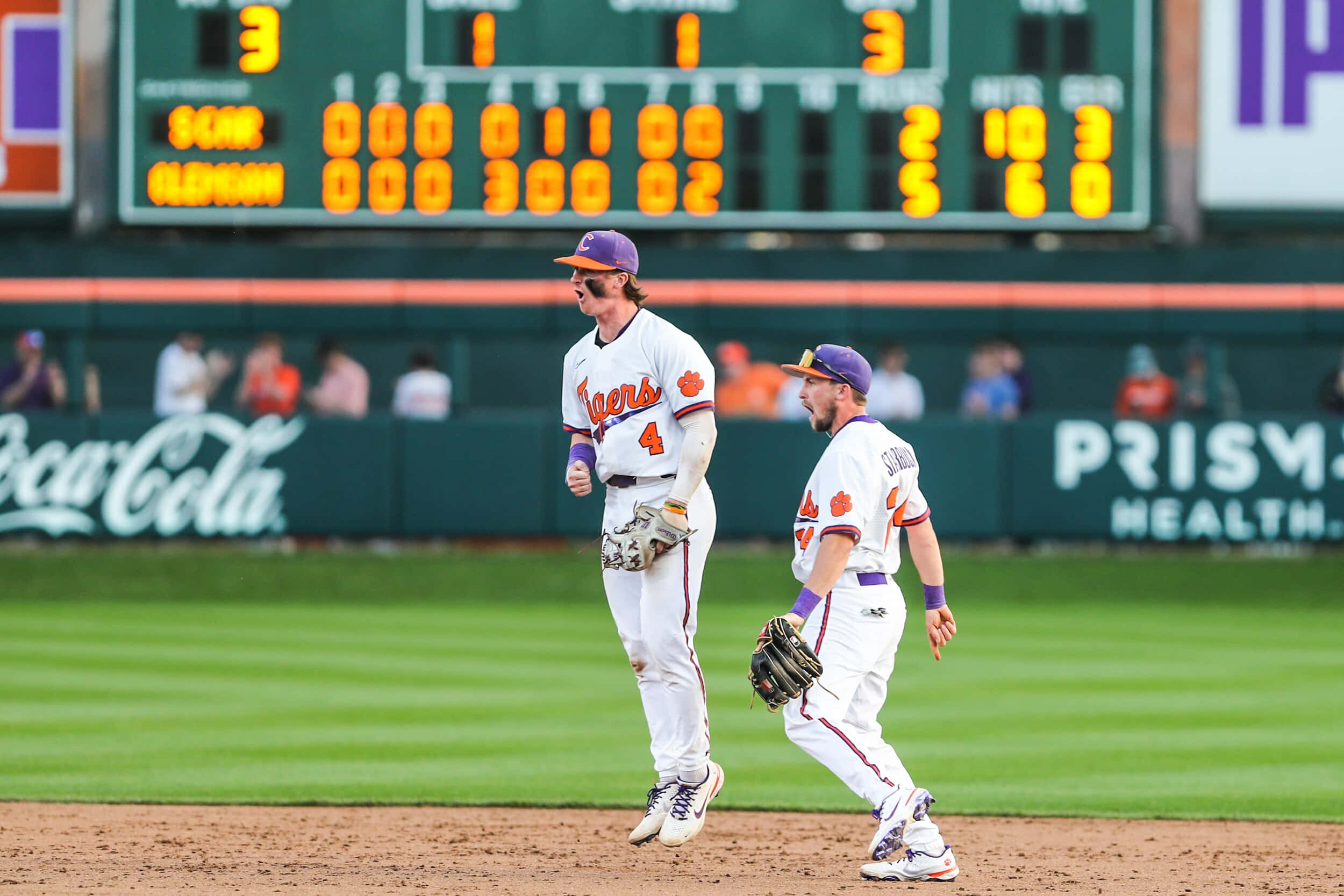 College Baseball 桌布 壁紙 — 兩名身穿白色球衣、戴著紫色帽子的棒球運動員，在陽光照耀的球場上走過，朝著顯示比賽統計數據的記分板前進。