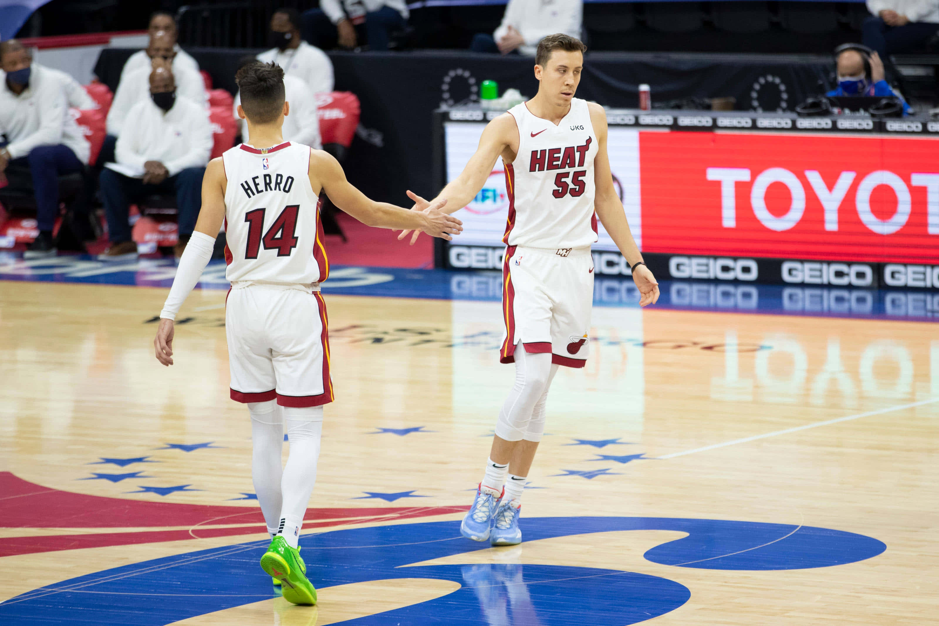 Tyler Herro 桌布 壁紙 — 兩名身穿白色球衣的邁阿密熱火籃球運動員在拋光木地板上握手，場地上可見球隊標誌和贊助商廣告。