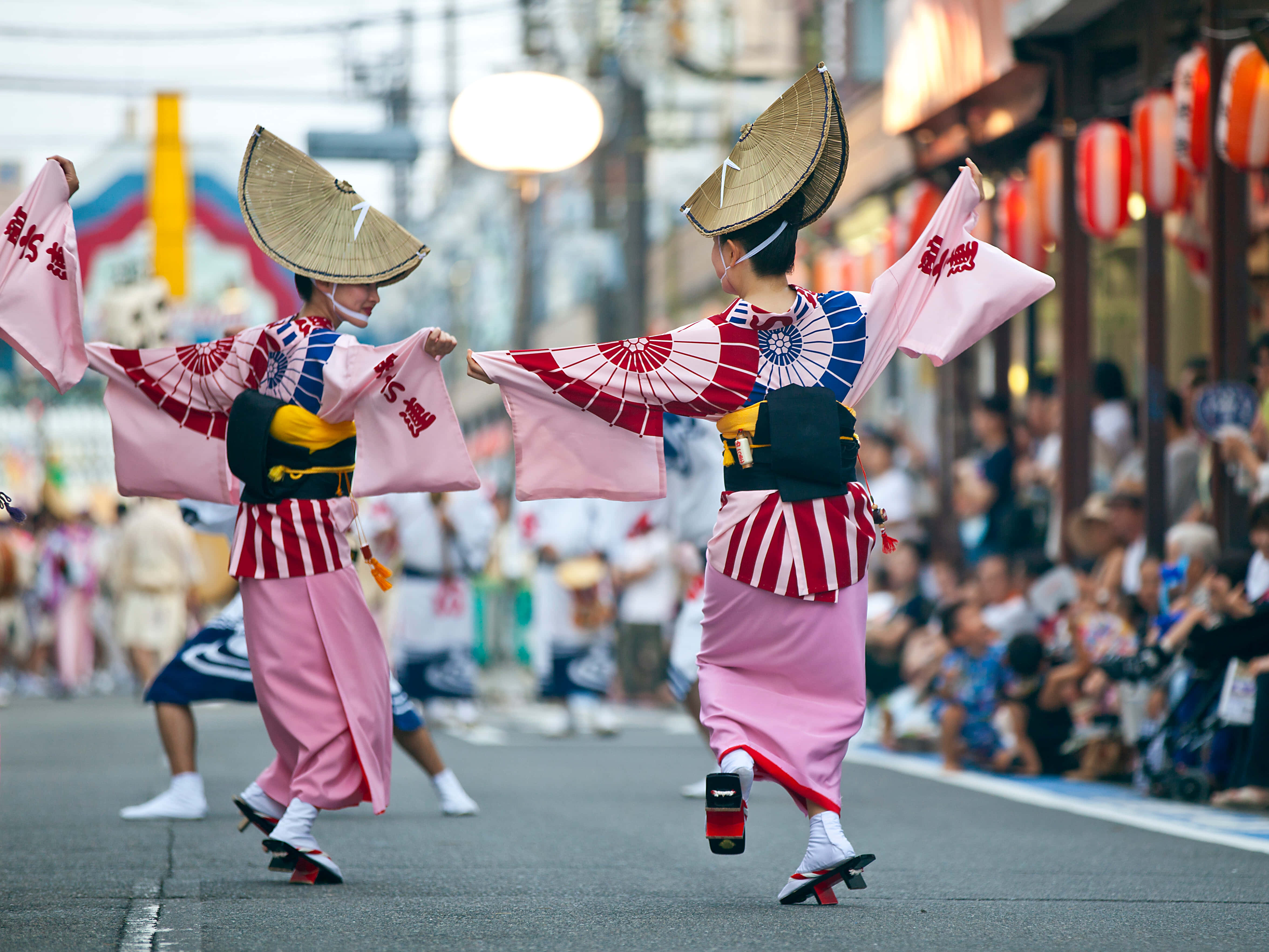 Japanese 桌布 壁紙 — 兩名身穿粉紅色和紅色傳統服飾、戴著草帽的舞者，在掛滿燈籠的熱鬧日本街道上充滿活力地表演。