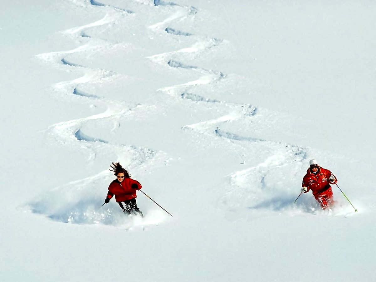 Skiing 桌布 壁紙 — 兩名身穿紅色外套的滑雪者在明亮的冬季斜坡上，在深粉雪中劃出動態的S形軌跡。