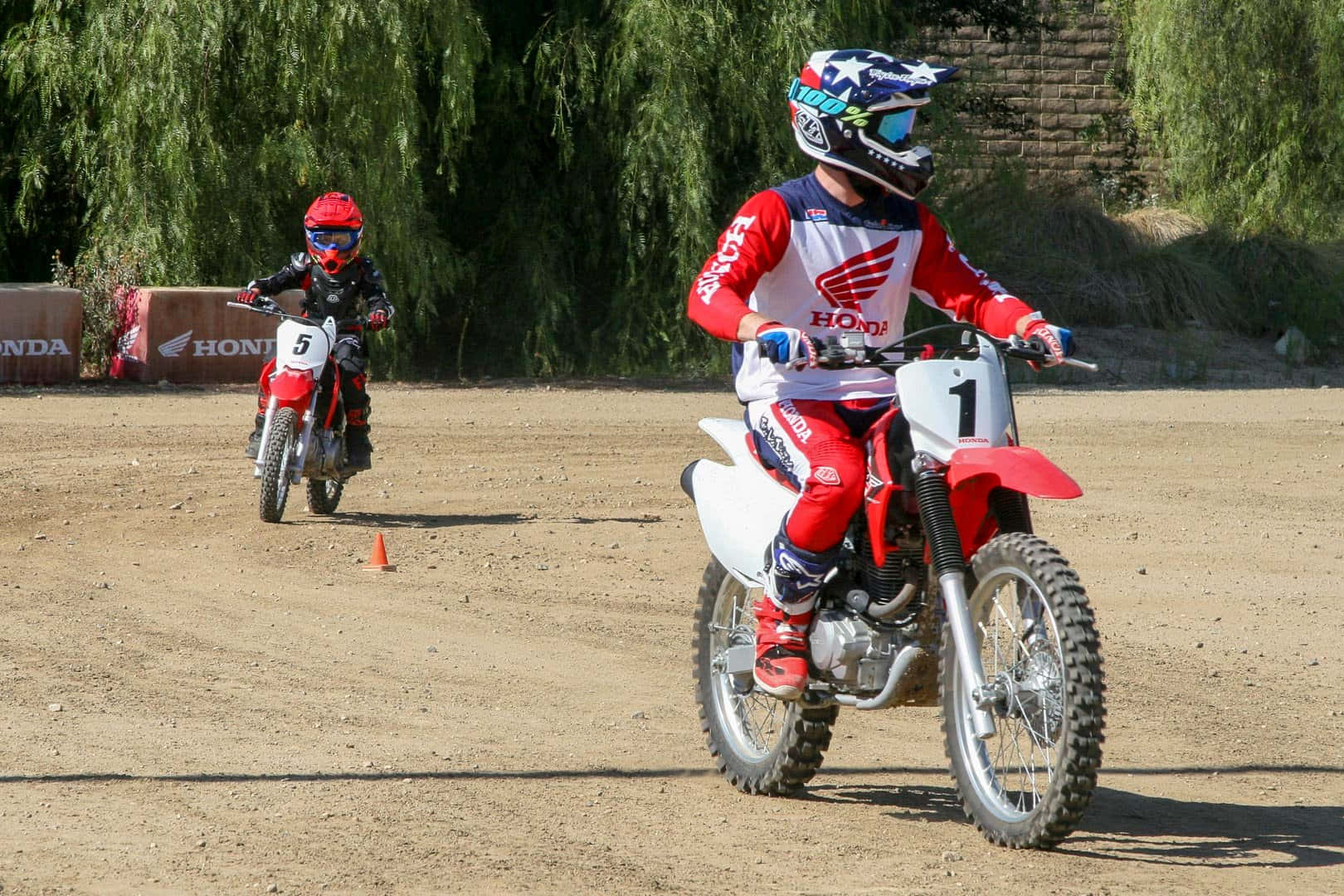 Dirtbike 桌布 壁紙 — 兩名身穿紅色裝備的年輕越野電單車手，在陽光明媚、塵土飛揚的賽道上駕駛紅色本田越野電單車，背景有綠樹。