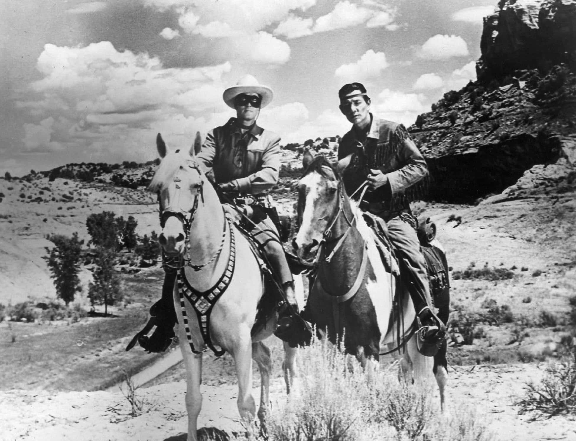 Black And White Film 桌布 壁紙 — 兩名身穿經典西部服裝的牛仔騎馬穿過陽光明媚的沙漠峽谷，兩旁是岩石峭壁和稀疏的植被。