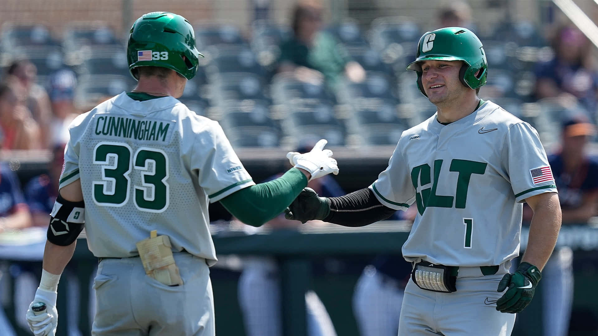 College Baseball 桌布 壁紙 — 兩名身穿綠色和白色球衣的棒球運動員在陽光明媚的球場上擊掌，背景是體育場座位。