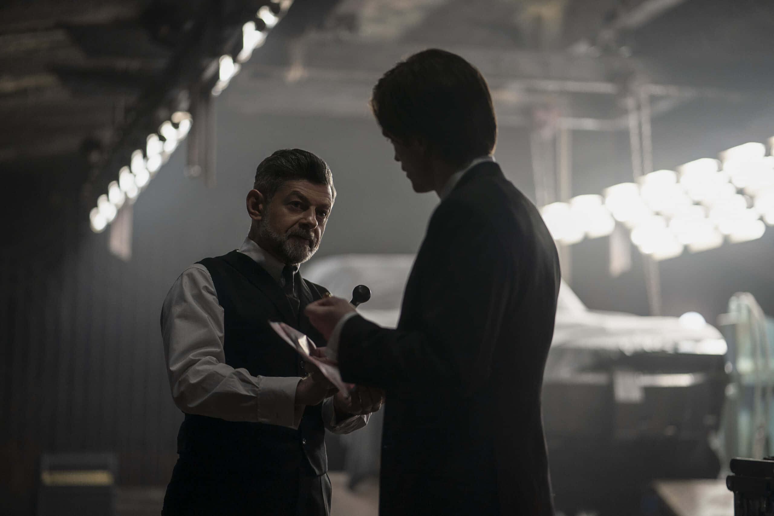 Alfred Pennyworth 桌布 壁紙 — 兩名身穿西裝的男子在昏暗的工業車庫中，在刺眼的頭頂燈光下交換文件。