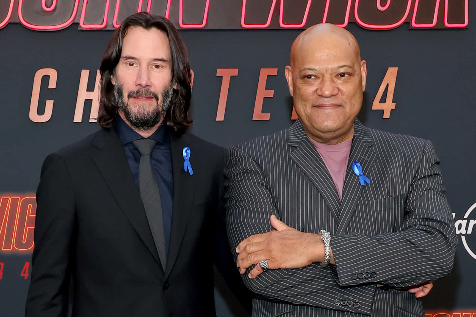 Keanu Reeves 桌布 壁紙 — 兩名身穿西裝的男子在電影首映禮上自信地站立，翻領上別著藍色絲帶。