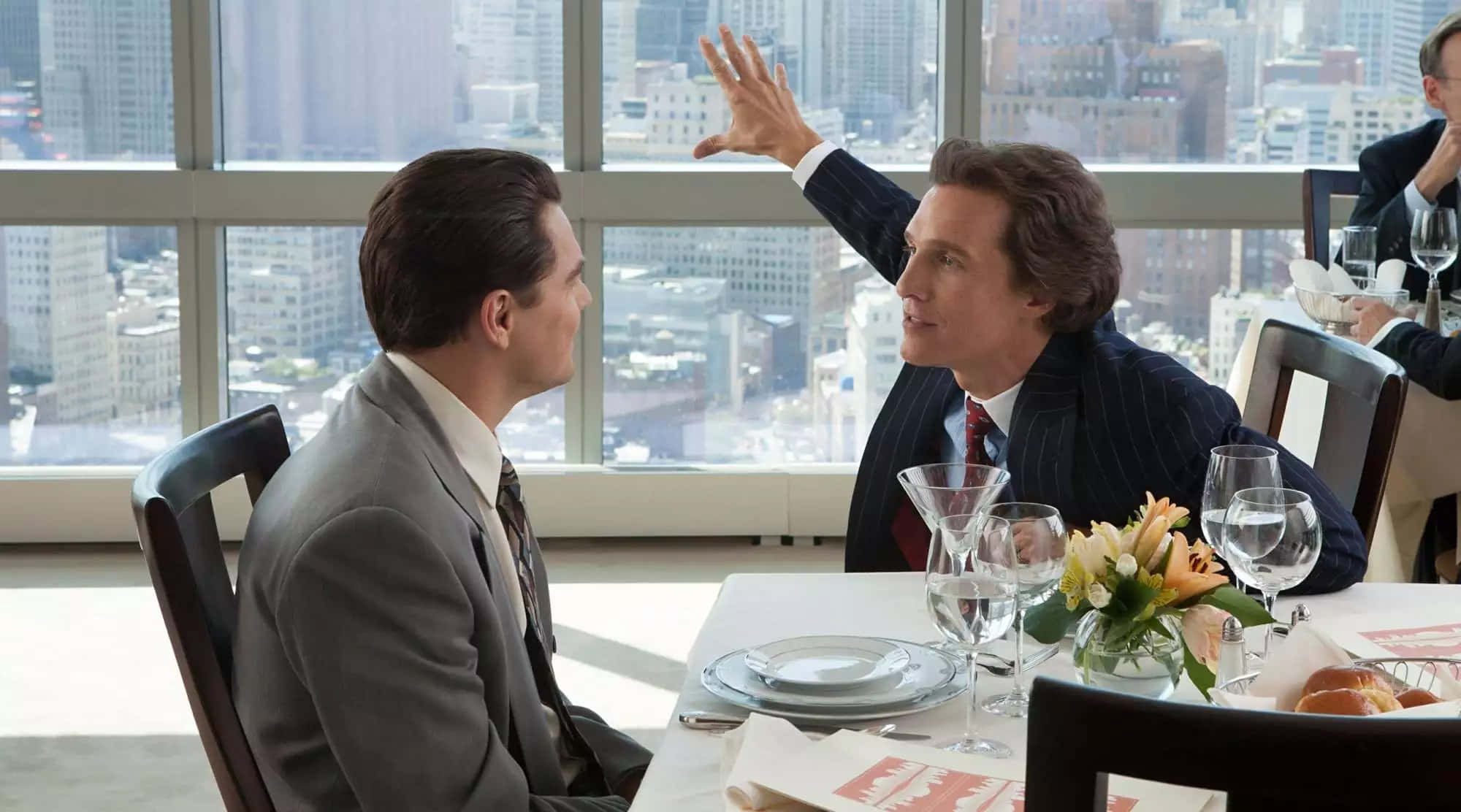 Wolf Of Wall Street 桌布 壁紙 — 兩名身穿西裝的男子在高層餐廳的餐桌旁進行緊張的對話，窗外是全景城市景觀。