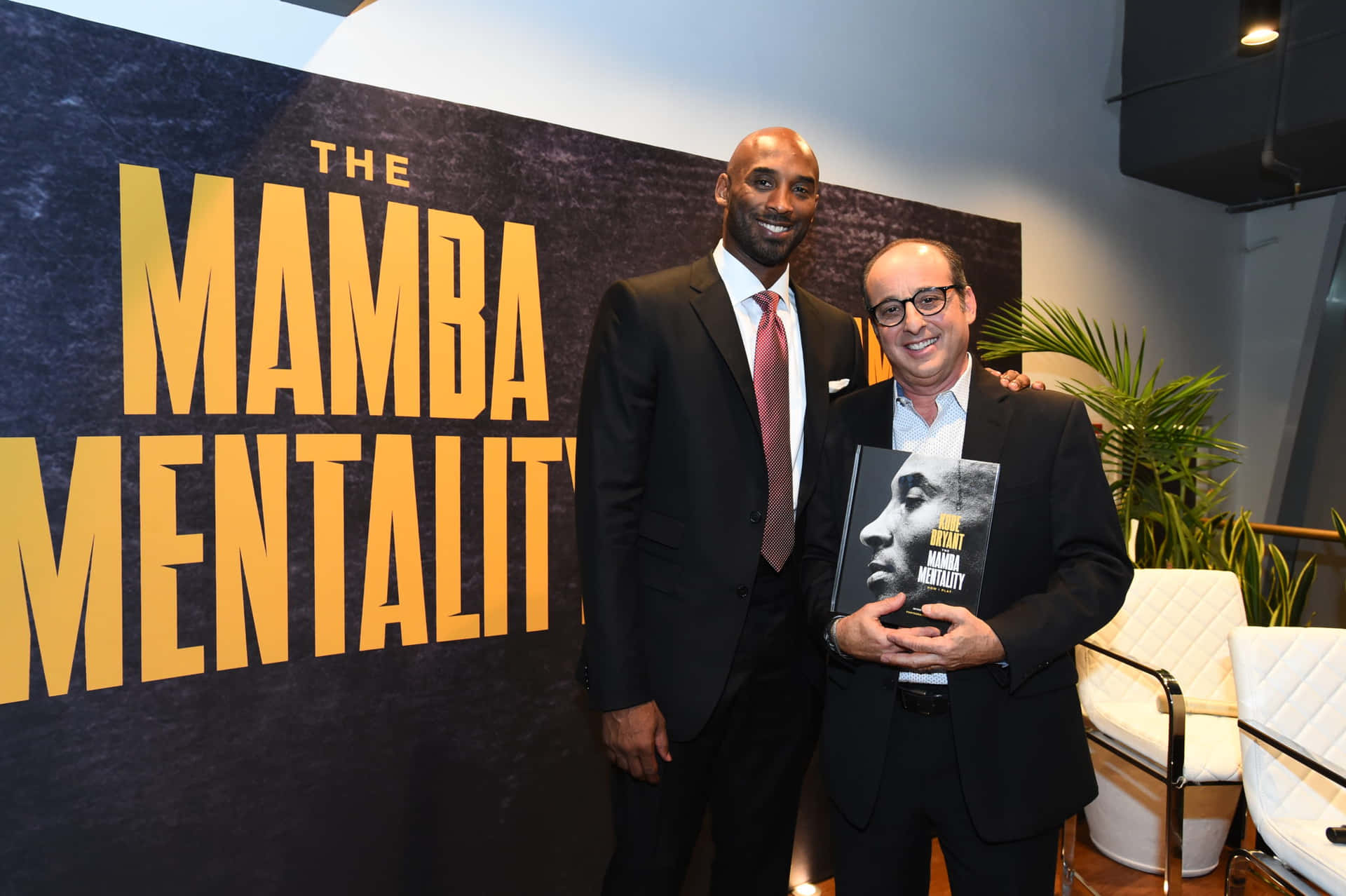 Mamba Mentality 桌布 壁紙 — 兩名身穿西裝的男子微笑著，手持名為《The Mamba Mentality》的書籍，在品牌背景前擺姿勢。