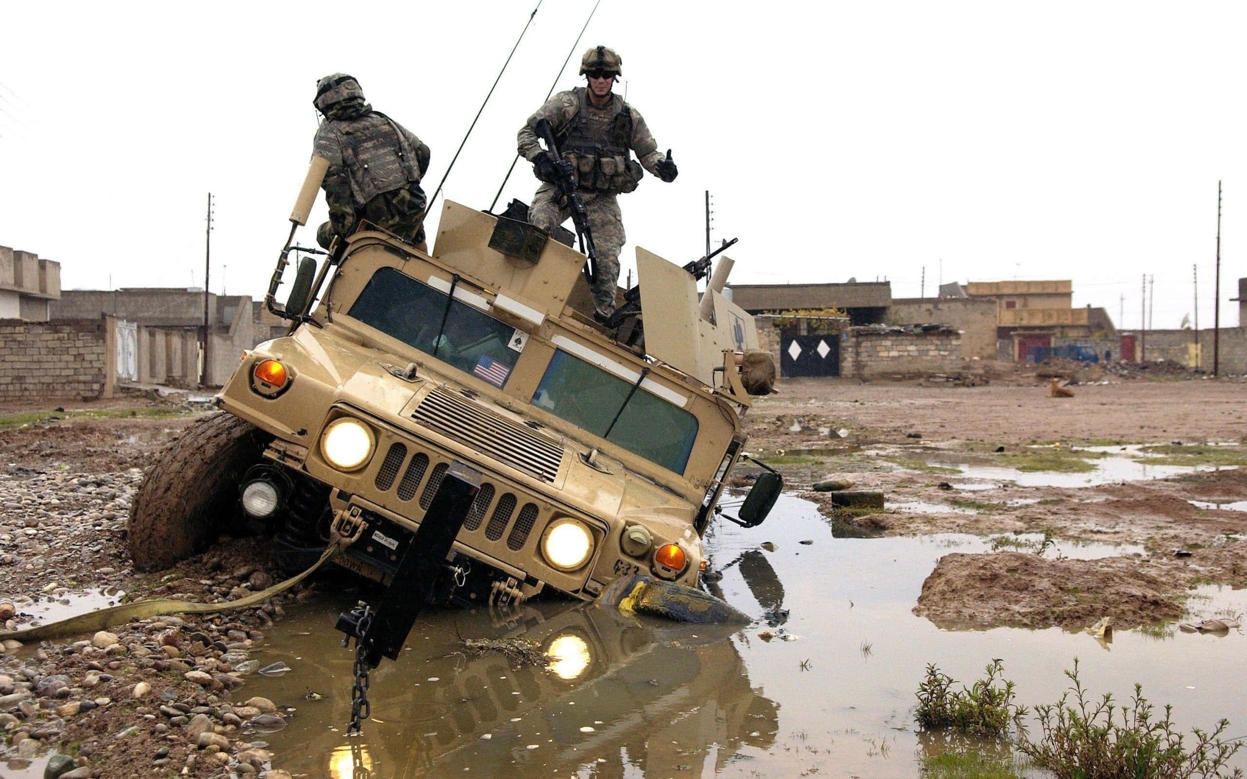 Army 桌布 壁紙 — 兩名身穿迷彩裝備的士兵站在一輛卡在泥水中的棕褐色軍用悍馬車頂上，附近是破舊的建築物。