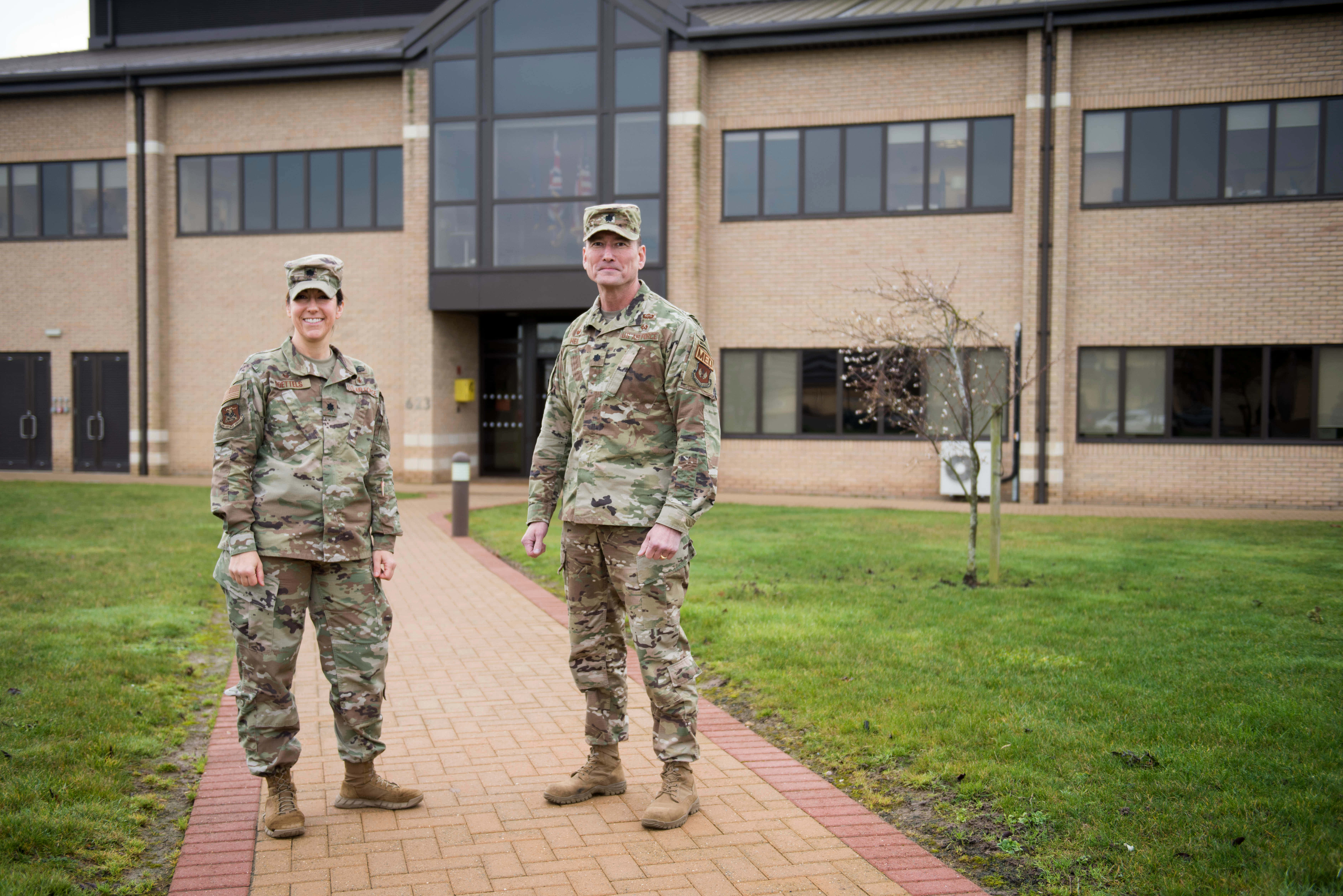 Air Force 桌布 壁紙 — 兩名身穿迷彩軍裝、面帶微笑的士兵站在磚砌人行道上，身後是一座帶有大窗戶的現代磚砌建築。
