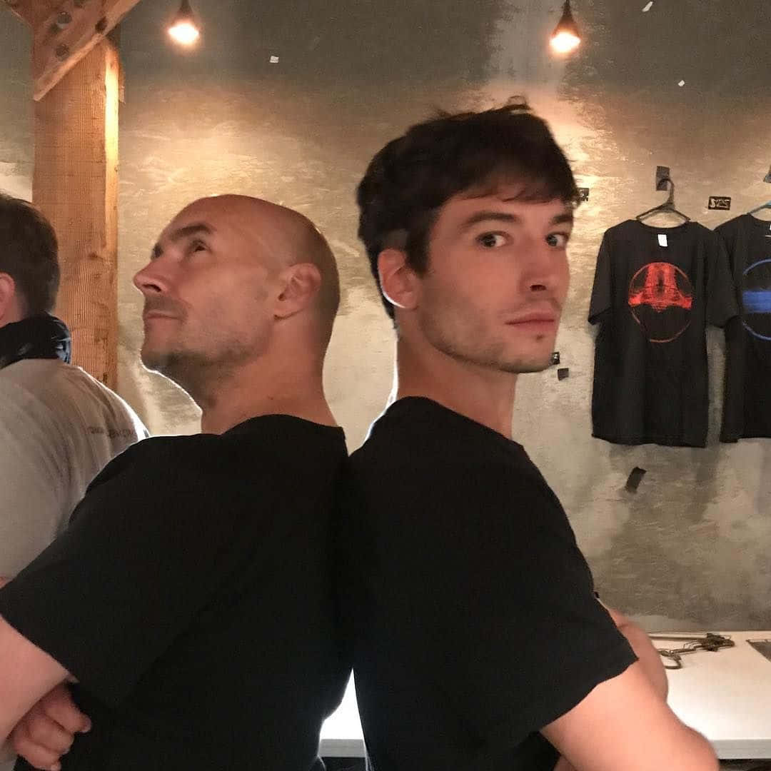 Grant Morrison 桌布 壁紙 — 兩名身穿黑色T恤的男子在一個光線溫暖的室內空間中背對背站立，周圍掛有襯衫並露出木樑。