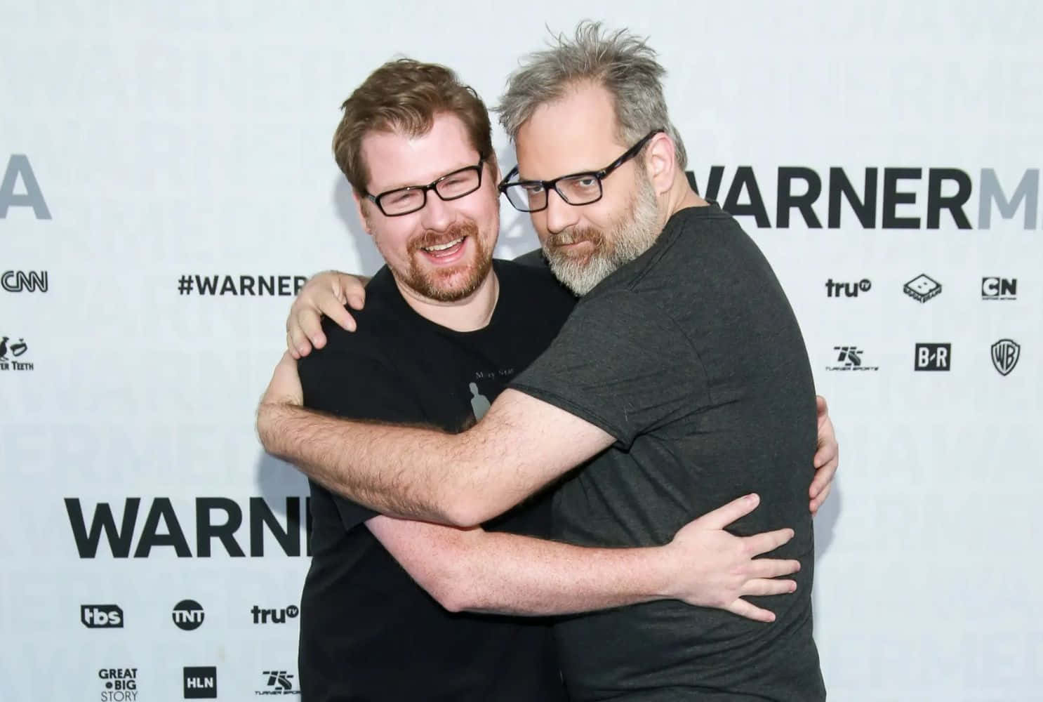 Justin Roiland 桌布 壁紙 — 兩名身穿黑色T恤的男子在帶有標誌的WarnerMedia活動背景前熱情擁抱。