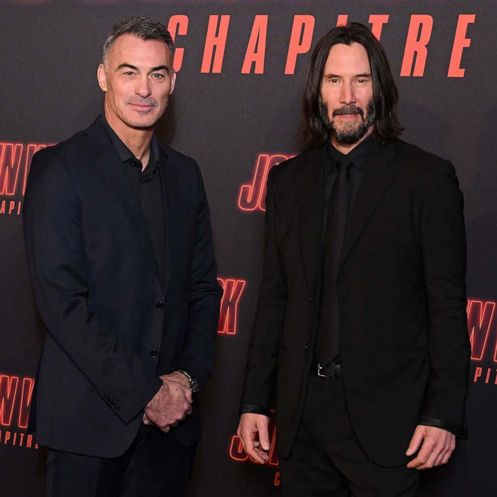 Keanu Reeves 桌布 壁紙 — 兩名身穿黑色西裝的男子在電影首映禮上自信地擺姿勢，身後有發光的紅色字母。