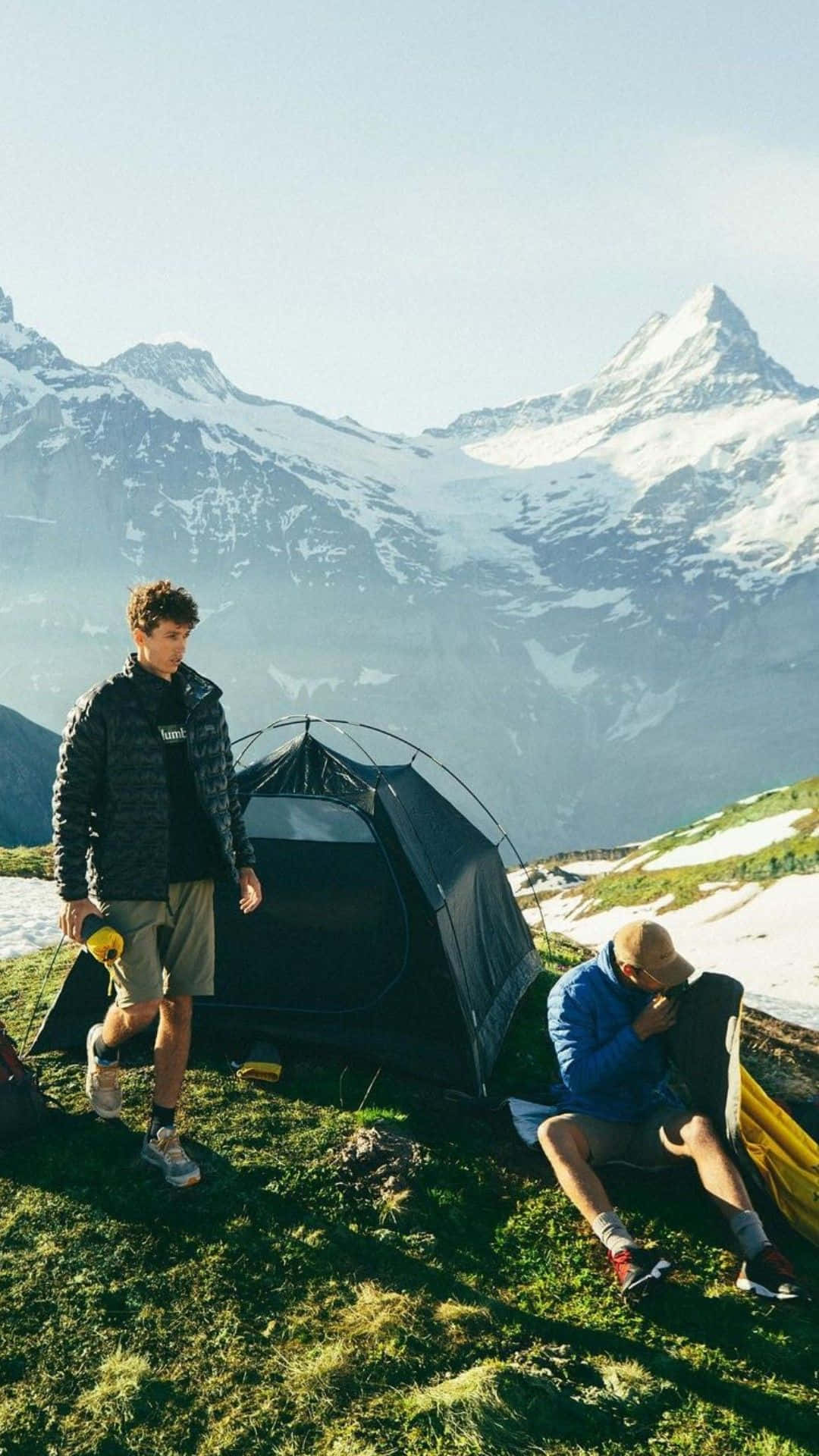Minimalist Camping 桌布 壁紙 — 兩名遠足人士在陽光照耀的草地上，靠近一個黑色帳篷休息，背景是白雪覆蓋的山峰高聳入雲。