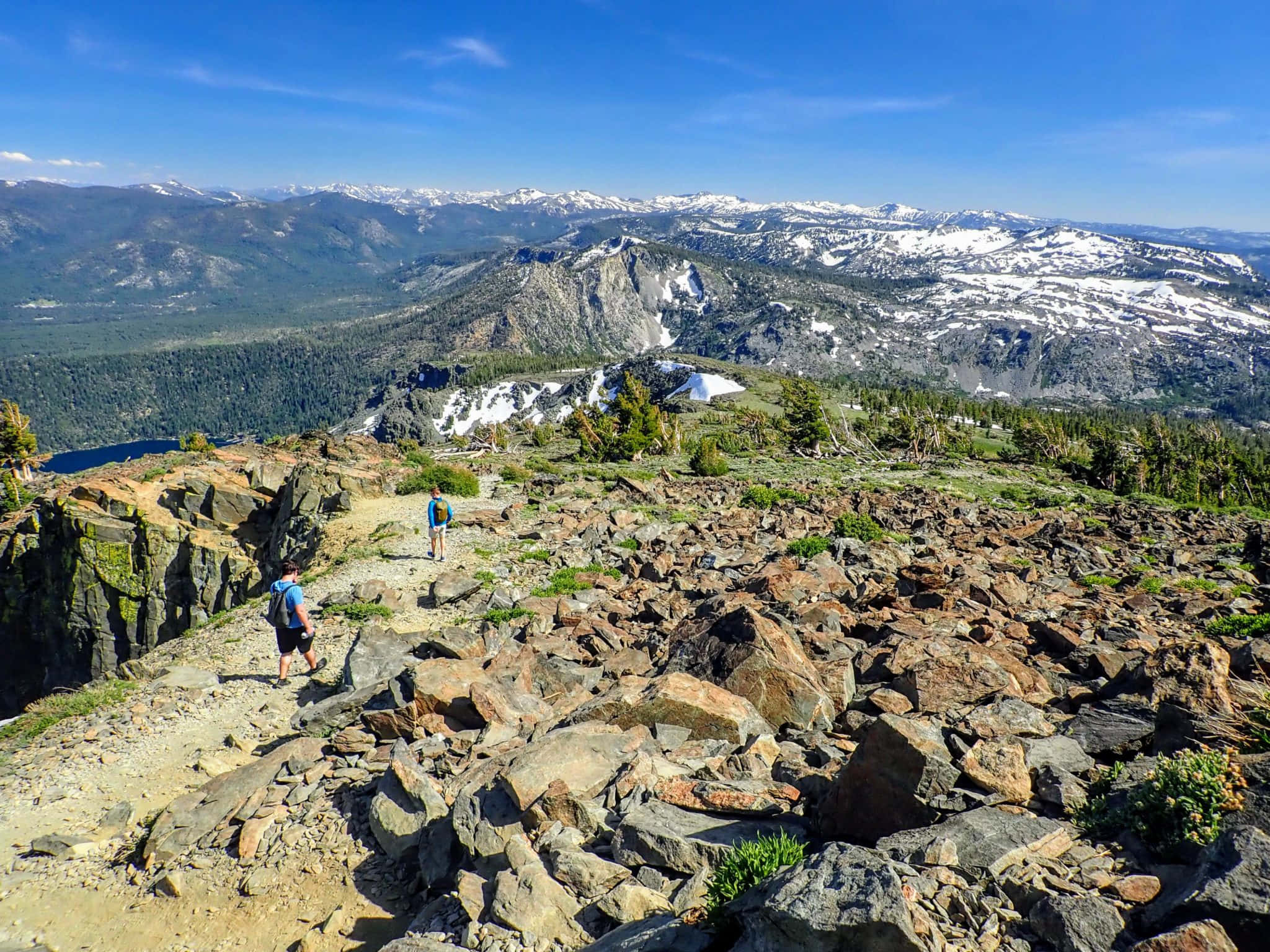 Hiking 桌布 壁紙 — 兩名遠足者穿越陽光明媚的岩石山脊，俯瞰白雪覆蓋的山峰和森林覆蓋的山谷，天空清澈湛藍。