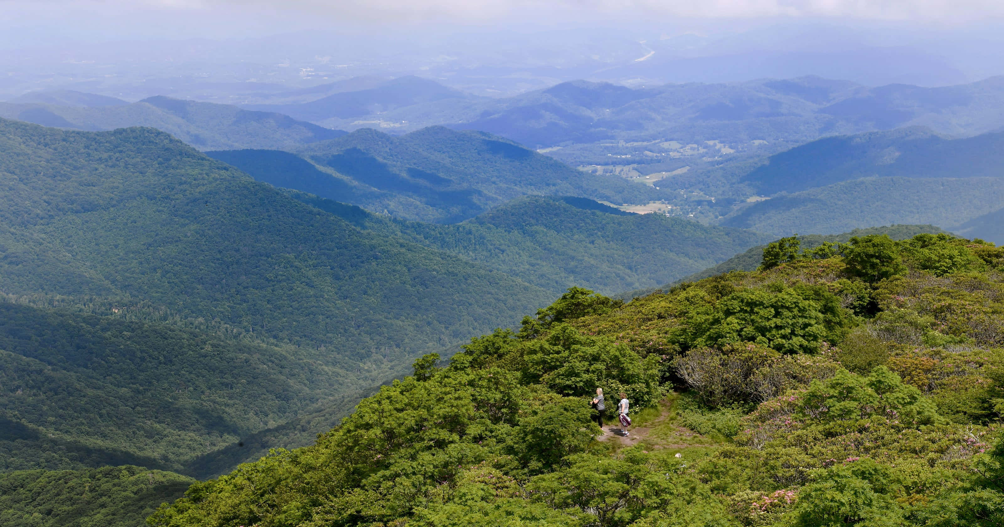 Blue Ridge Parkway 桌布 壁紙 — 兩名遠足者穿越陽光普照的山脊，俯瞰在朦朧天空下無盡的綠色山谷和遠處的藍色山峰。
