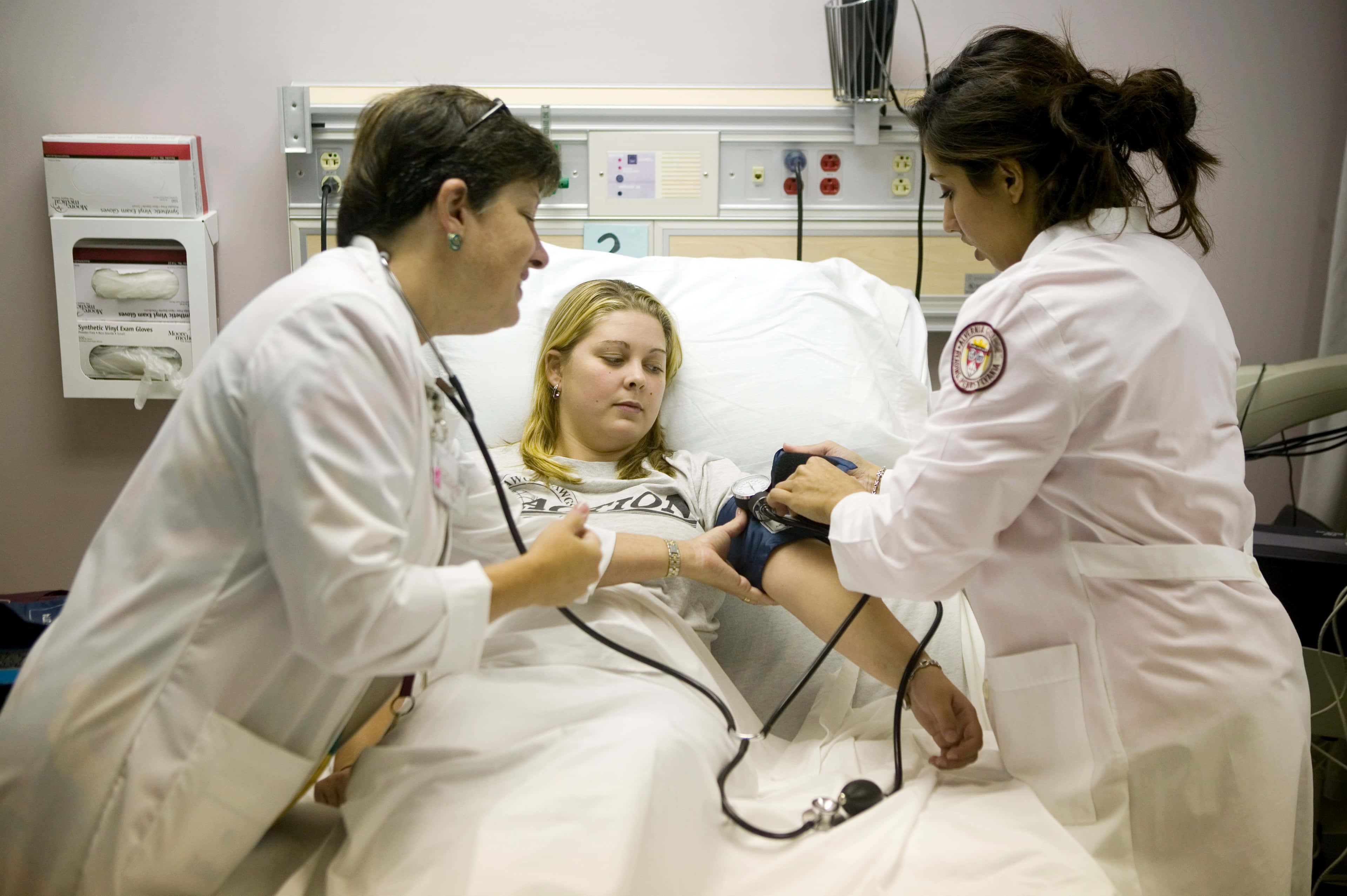 Nursing 桌布 壁紙 — 兩名醫護人員正在用血壓計袖帶和聽診器照顧病床上的病人。
