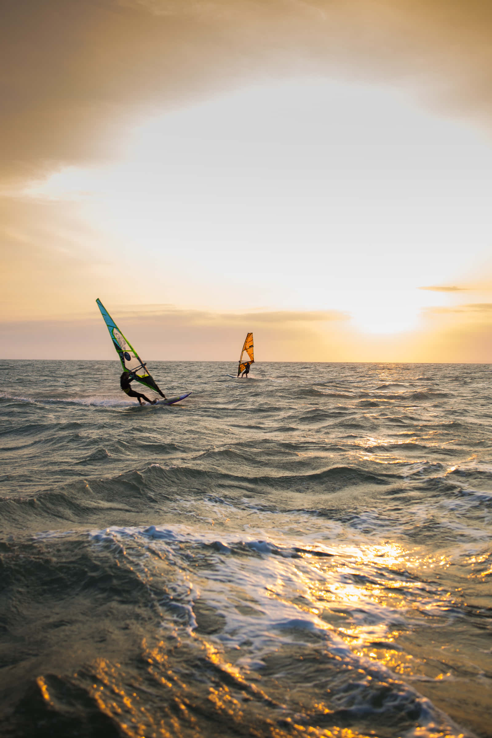 Beach Water Sports 桌布 壁紙 — 兩名風帆衝浪者在日落時分，在戲劇性的天空背景下，滑過金黃色的海浪。