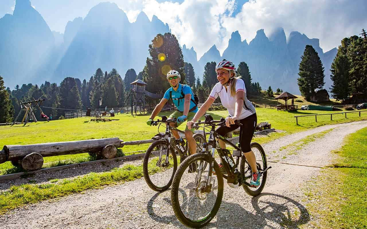 Electric Bike 桌布 壁紙 — 兩名騎單車人士在陽光明媚的碎石小徑上愉快地踩著踏板，穿過一片茂密的綠色草地，背景是高聳的鋸齒狀山脈。