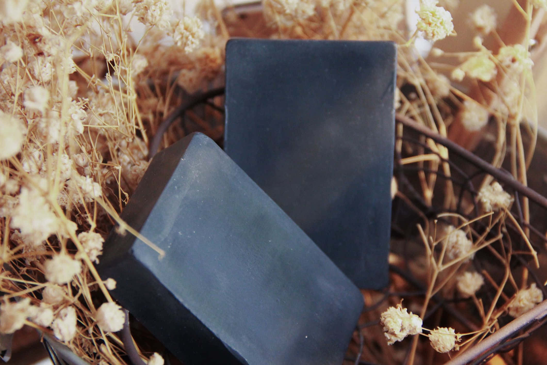 Black Soap 桌布 壁紙 — 兩塊啞光深藍色肥皂放在一個鐵絲籃中，周圍環繞著乾燥的滿天星花朵。