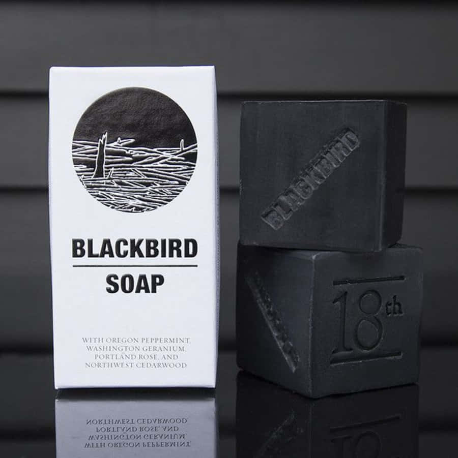 Black Soap 桌布 壁紙 — 兩塊啞光黑色肥皂疊放在一個極簡白色盒子旁邊，盒子上標有 Blackbird Soap，置於反光的深色表面上。